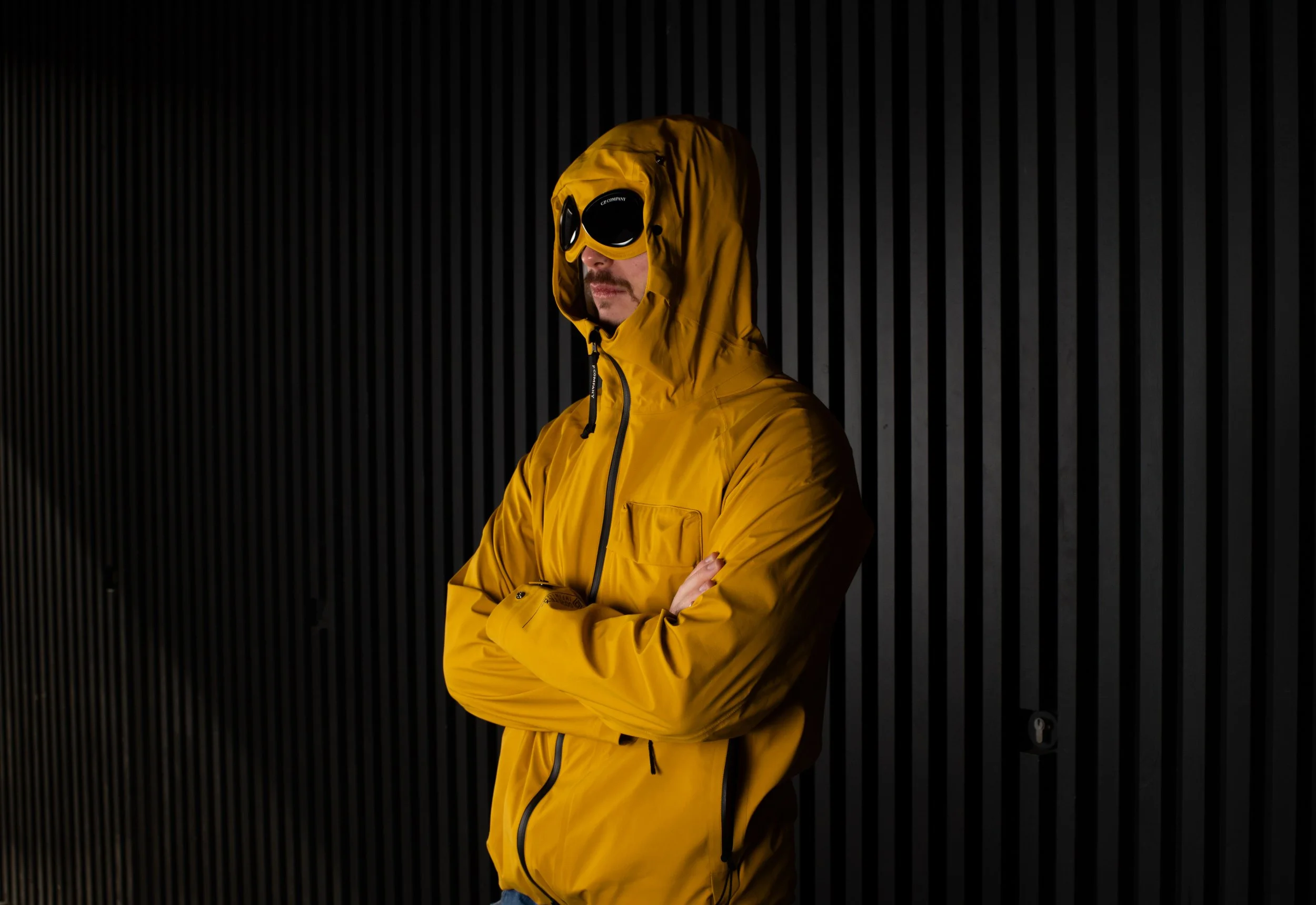 Man in a yellow jacket with hood and goggles stands confidently against a dark, striped background. The image conveys a sense of mystery and adventure.