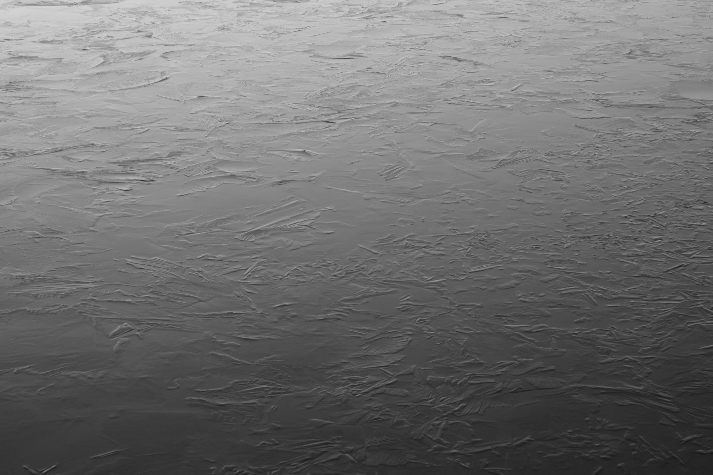 A monochrome image of a frozen surface, displaying intricate ice patterns and textures. The smooth, glass-like texture conveys a serene, cold atmosphere.