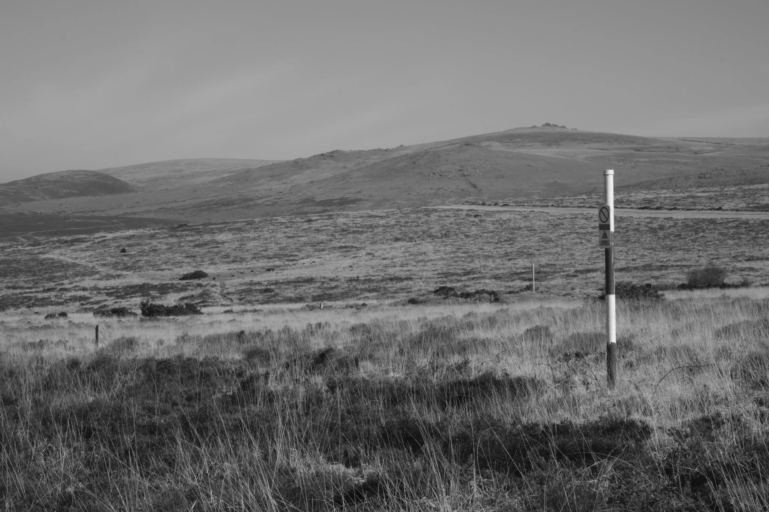 Open landscape with grassy fields and rolling hills in the background, a single striped post in the foreground, and a cloudy sky.