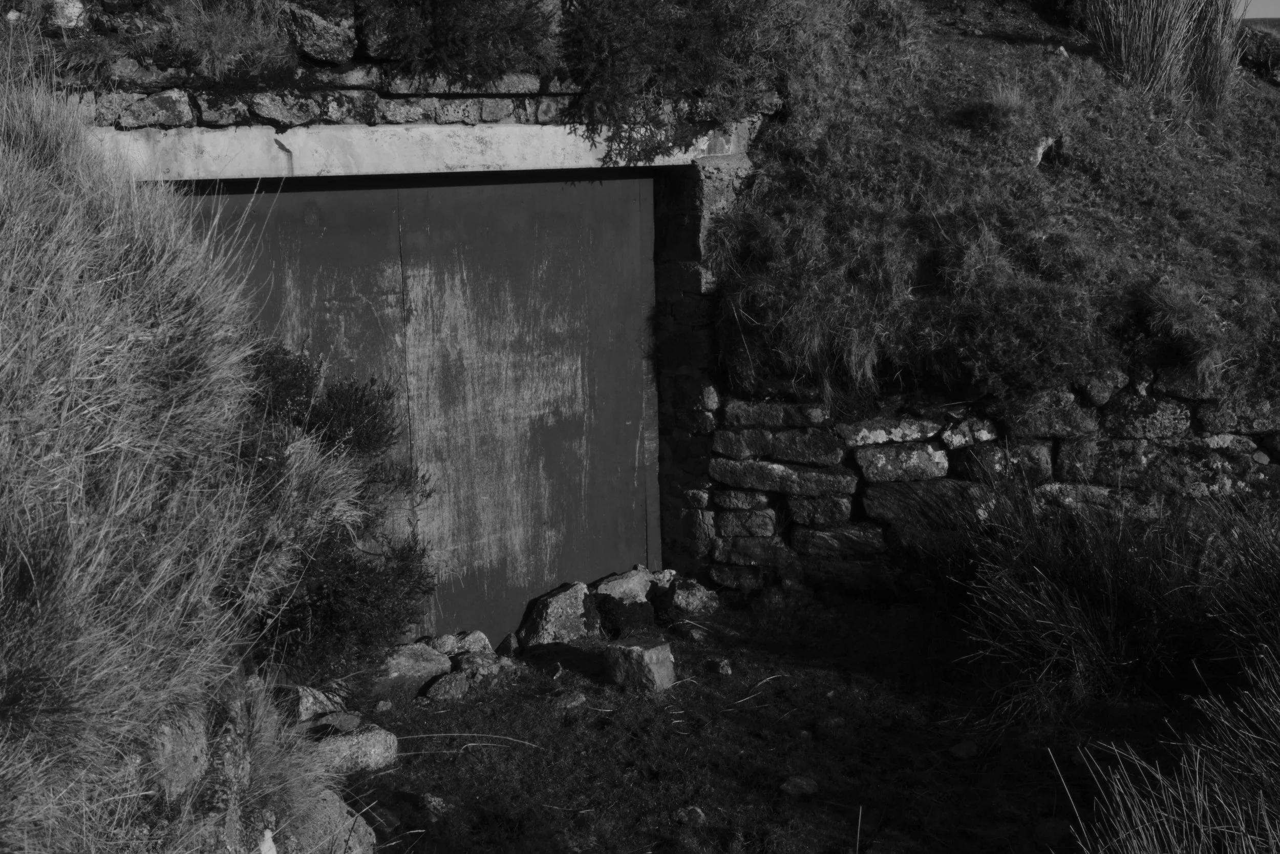 An old stone bunker door surrounded by rugged greenery and rocky terrain; the monochrome setting evokes a sense of mystery and abandonment.