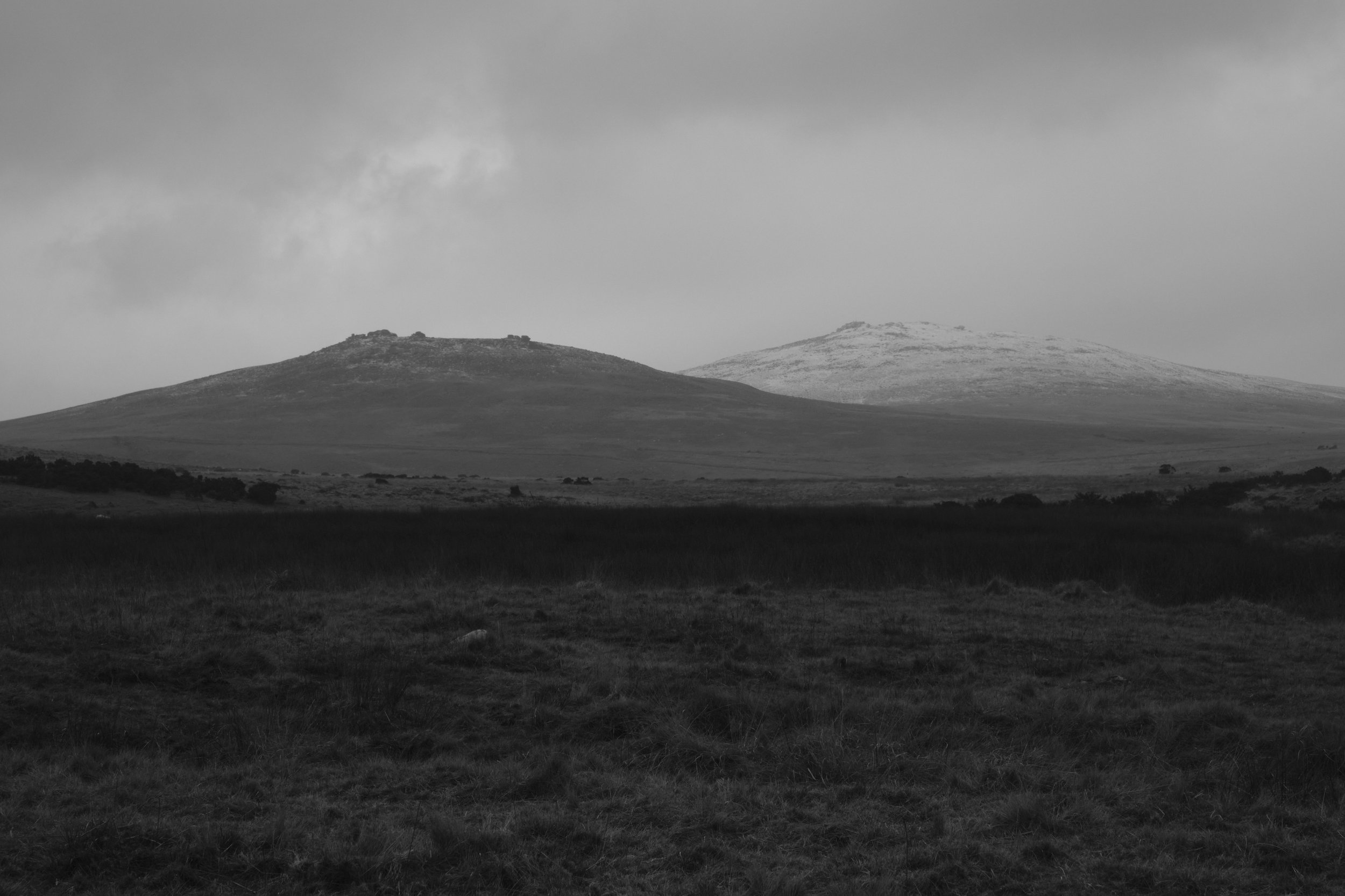 Monochrome landscape of two hills, with a light dusting of snow on the right hill under an overcast sky, conveying a serene, chilly atmosphere.