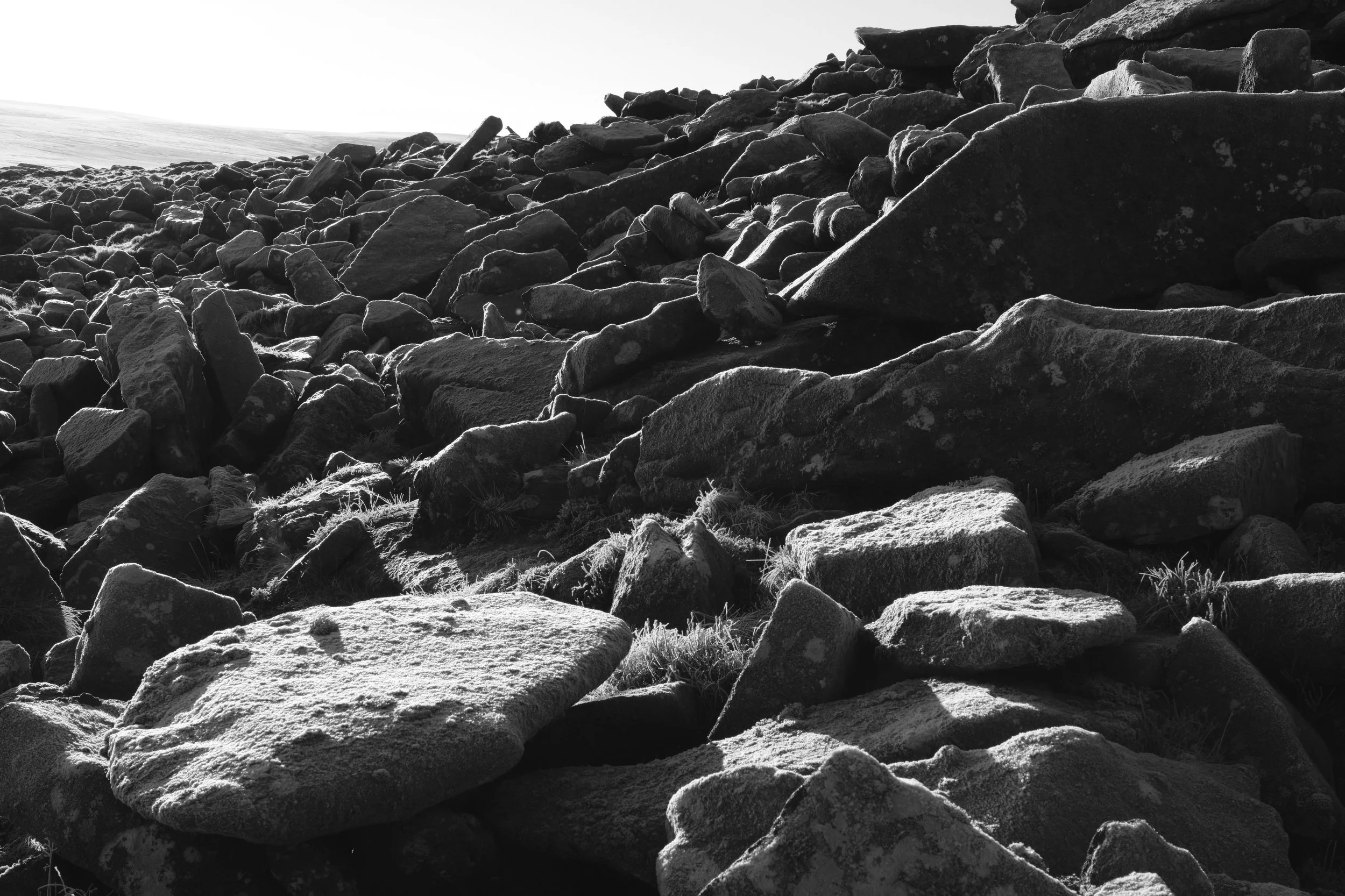 Black and white image of rough, sunlit rocks covering a hillside. The jagged terrain creates a stark, rugged landscape under a bright sky.