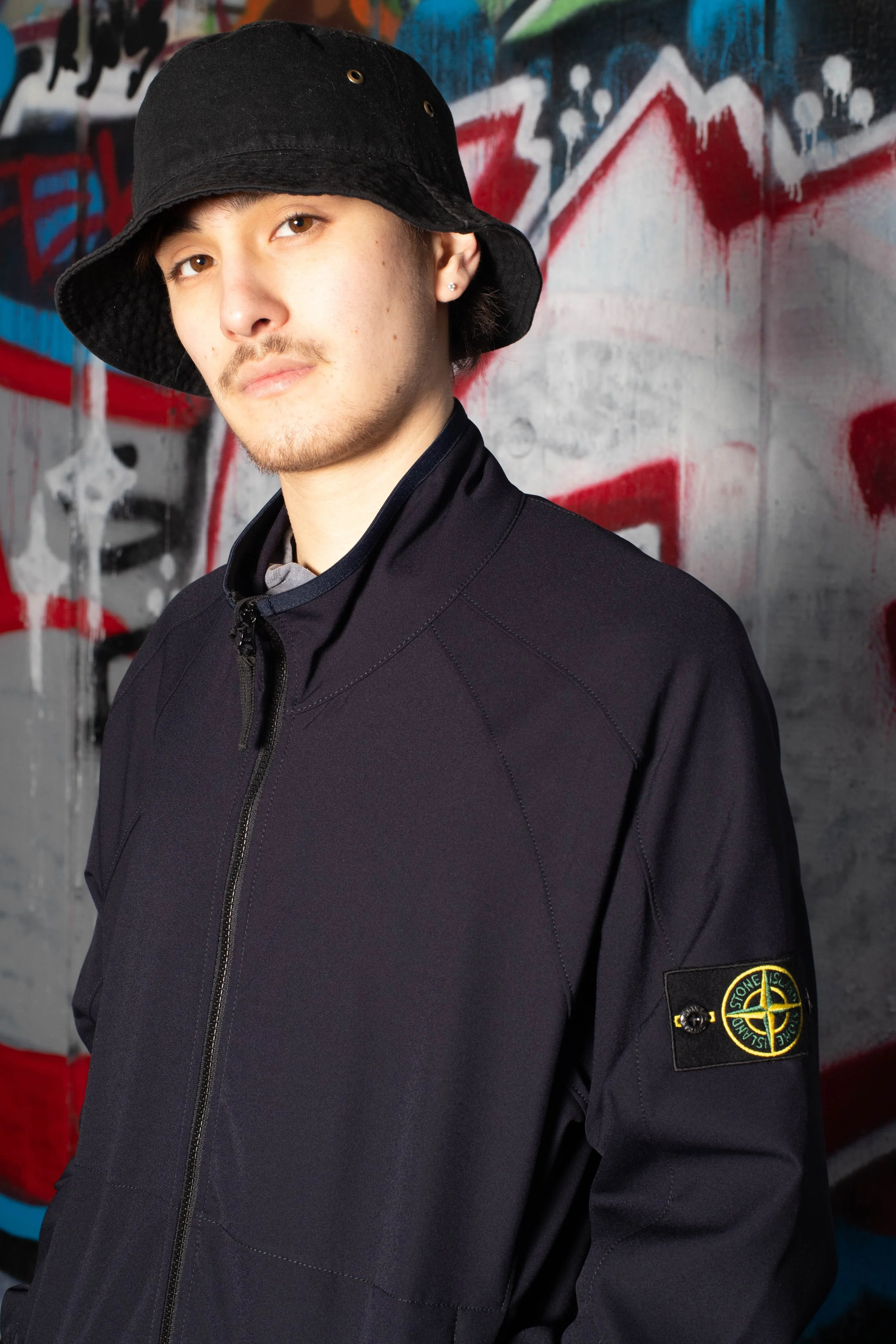 A person wearing a black bucket hat and navy jacket stands confidently against a vibrant graffiti wall. The mood is urban and stylish.