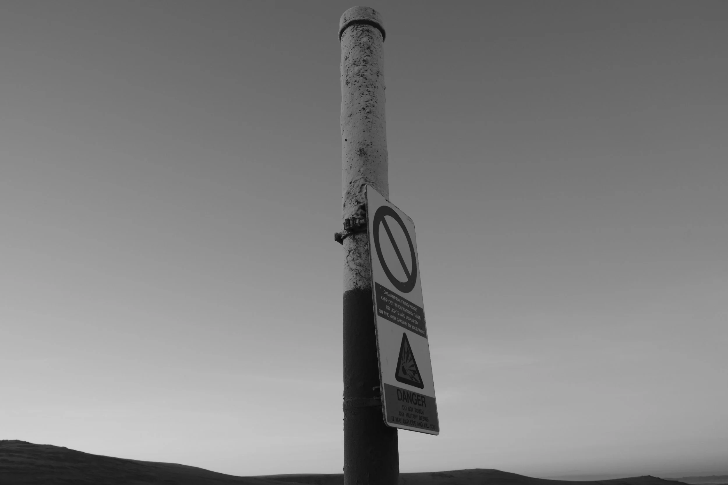 A black and white image of a tall, weathered pole with an attached no parking sign and faded caution symbol, set against a clear sky, evoking solitude.