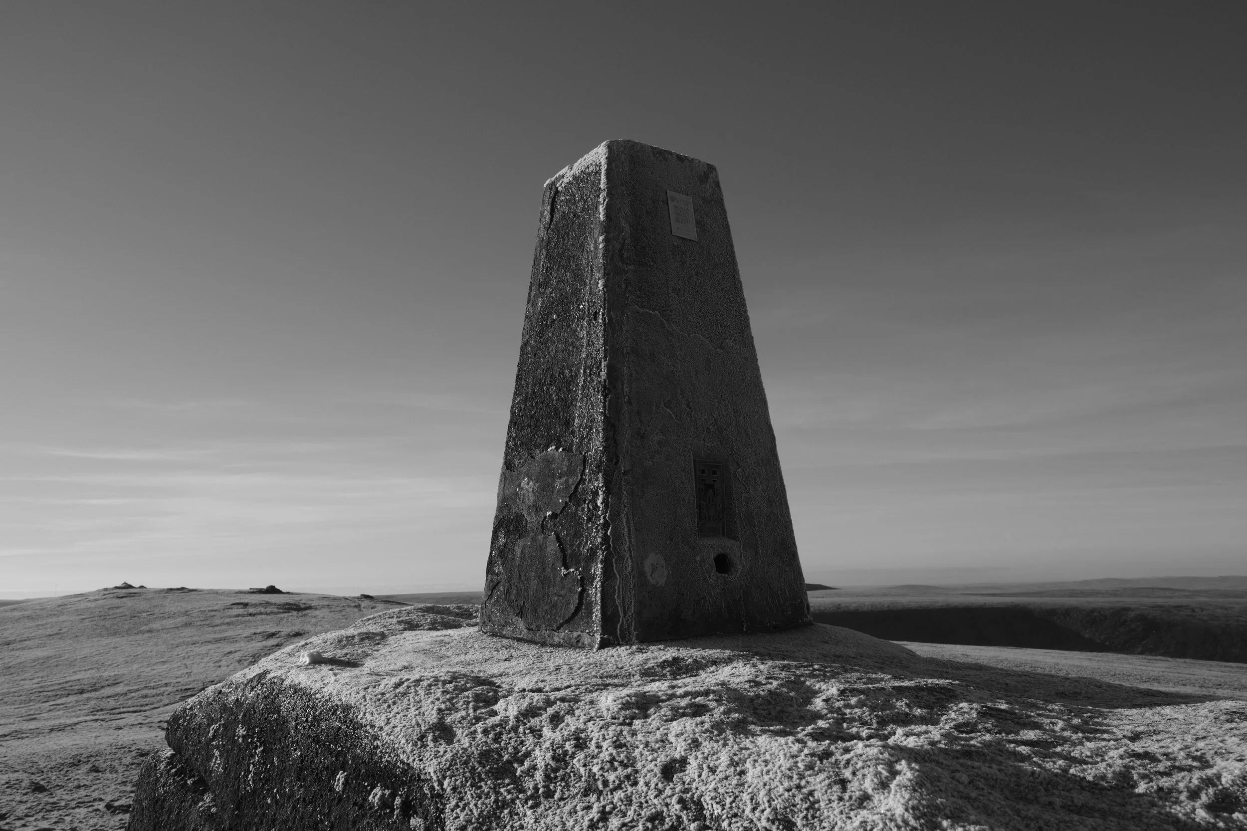 A weathered stone triangulation pillar atop a hill under clear skies in grayscale, conveying a sense of solitude and timelessness.