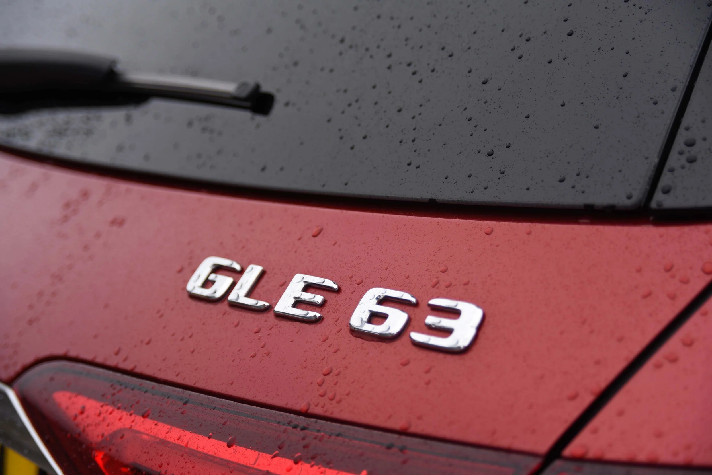 Close-up of a red car with water droplets, showing the chrome badge "GLE 63" on the rear.