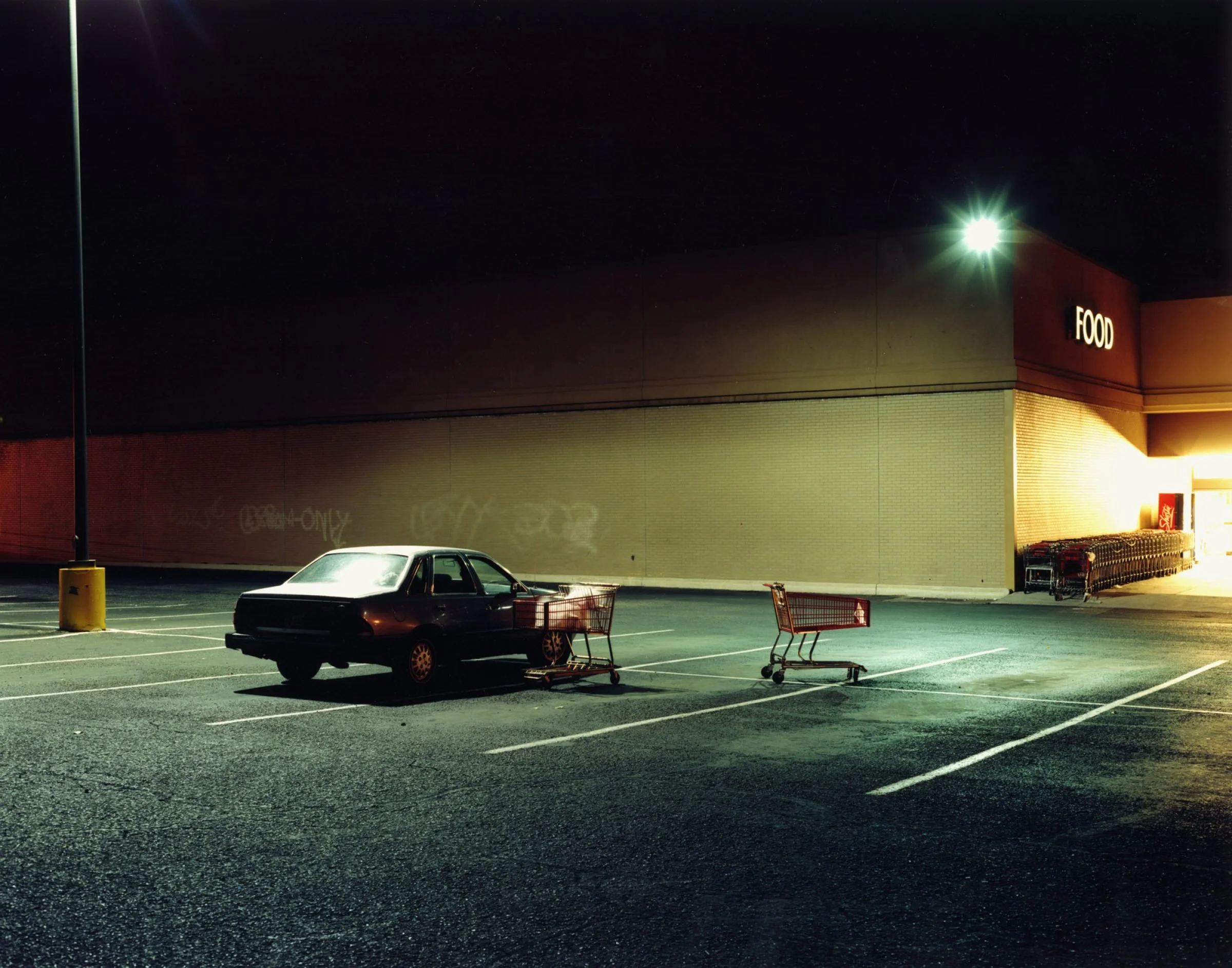 Colour photograph from Nadav Kander's God's Country depicting two shopping trollies next to a car parked in a supermarket car park lit by a single light.