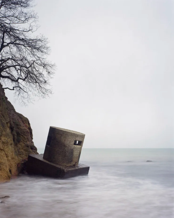 An old pillbox leans over the sea in a colour photograph with a tree visible slightly above it.