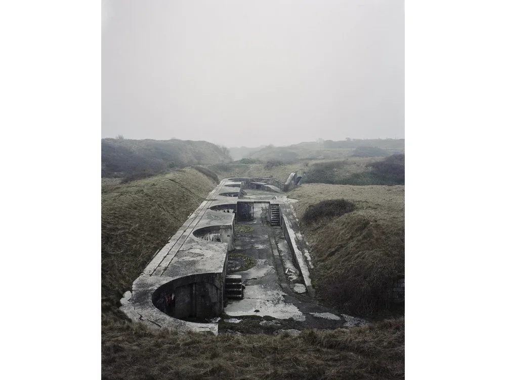 Colour photograph of an old grey military complex against a green and hilly backdrop in Dorset, England.