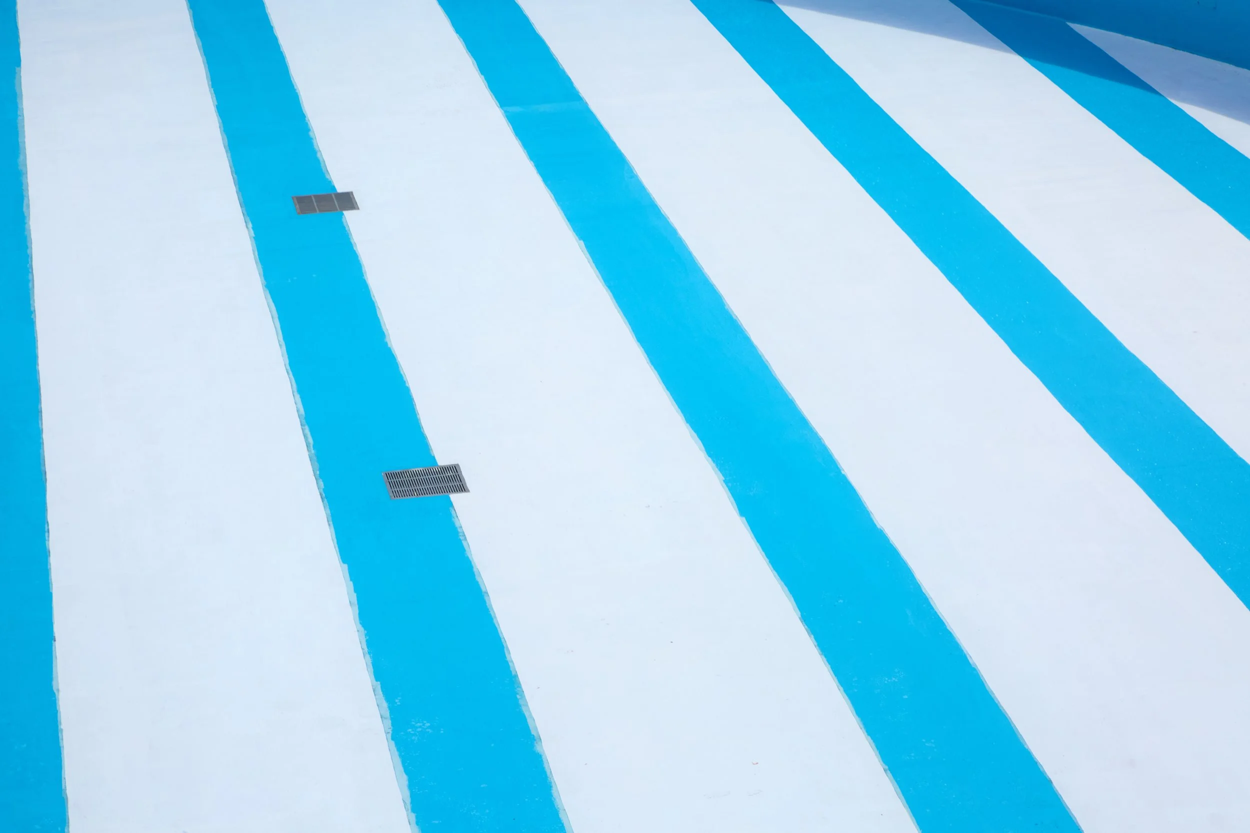 Empty swimming pool with alternating blue and white stripes, creating a bold pattern. Two small metal drains are visible, conveying minimalism and symmetry.