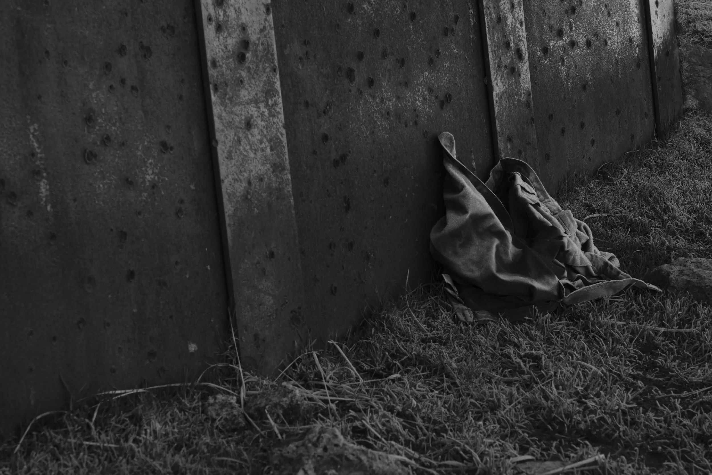 A weathered metal wall with bullet holes stands on grass. A crumpled, abandoned jacket lies against it, evoking a somber, desolate mood.