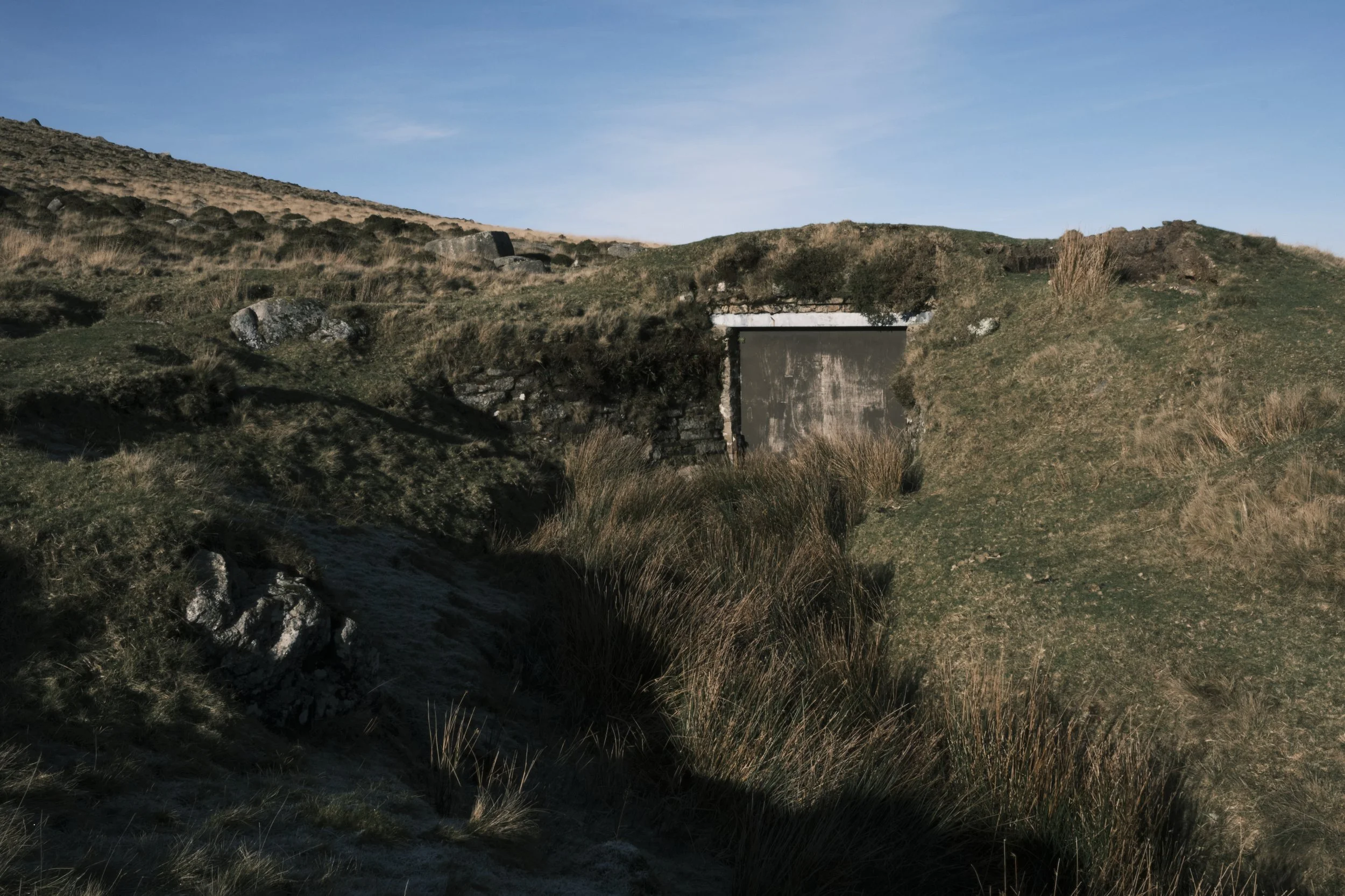 A concrete shed is built into a grassy hill with dry grass and rocks surrounding it. The sky is blue, and the scene conveys isolation and calmness.