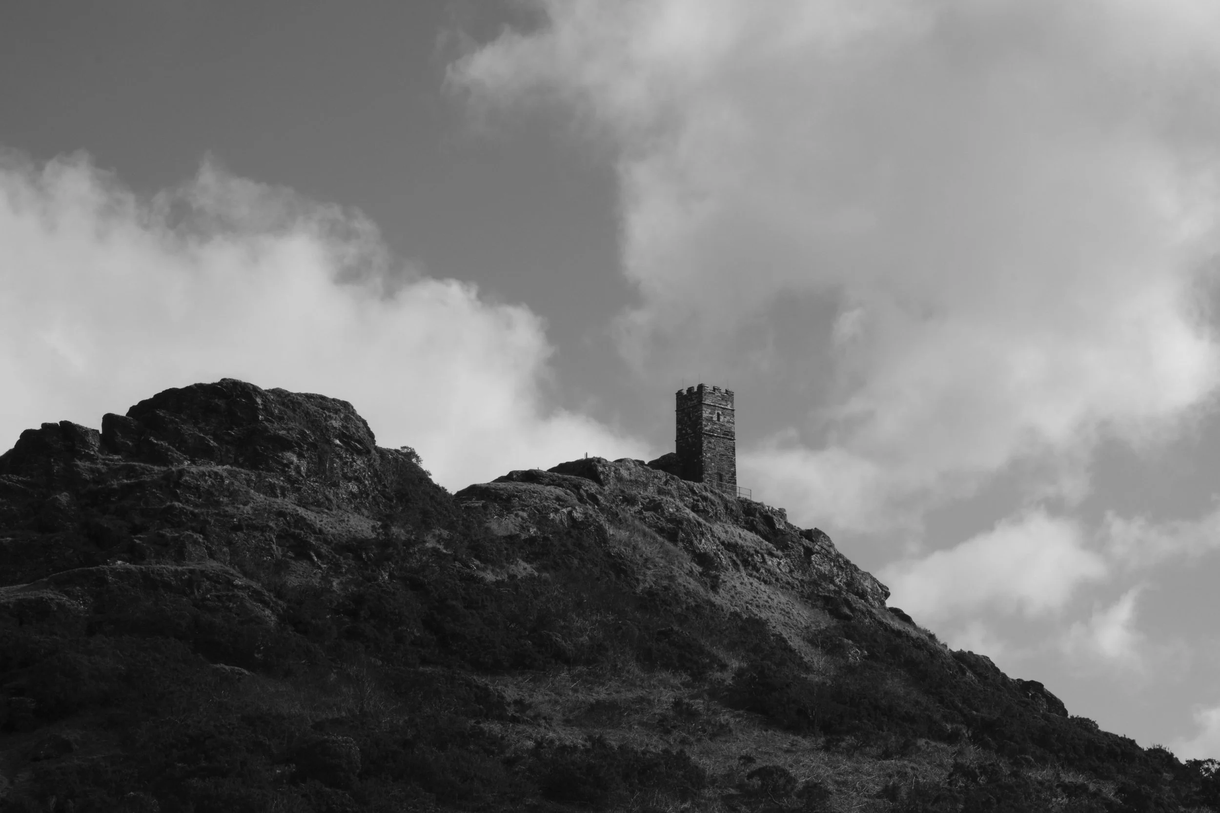 A black and white photo of a lone stone tower perched atop a rocky hill under a partly cloudy sky. The scene feels solitary and timeless.