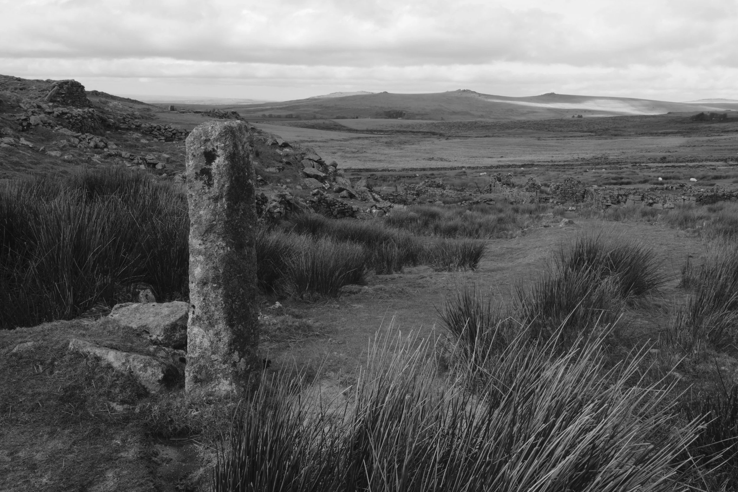 A black and white photograph of a post overlooking a landscape of overgrown grass and the tors of Dartmoor National Park with Foggintor Quarry visible on the left side of the image.