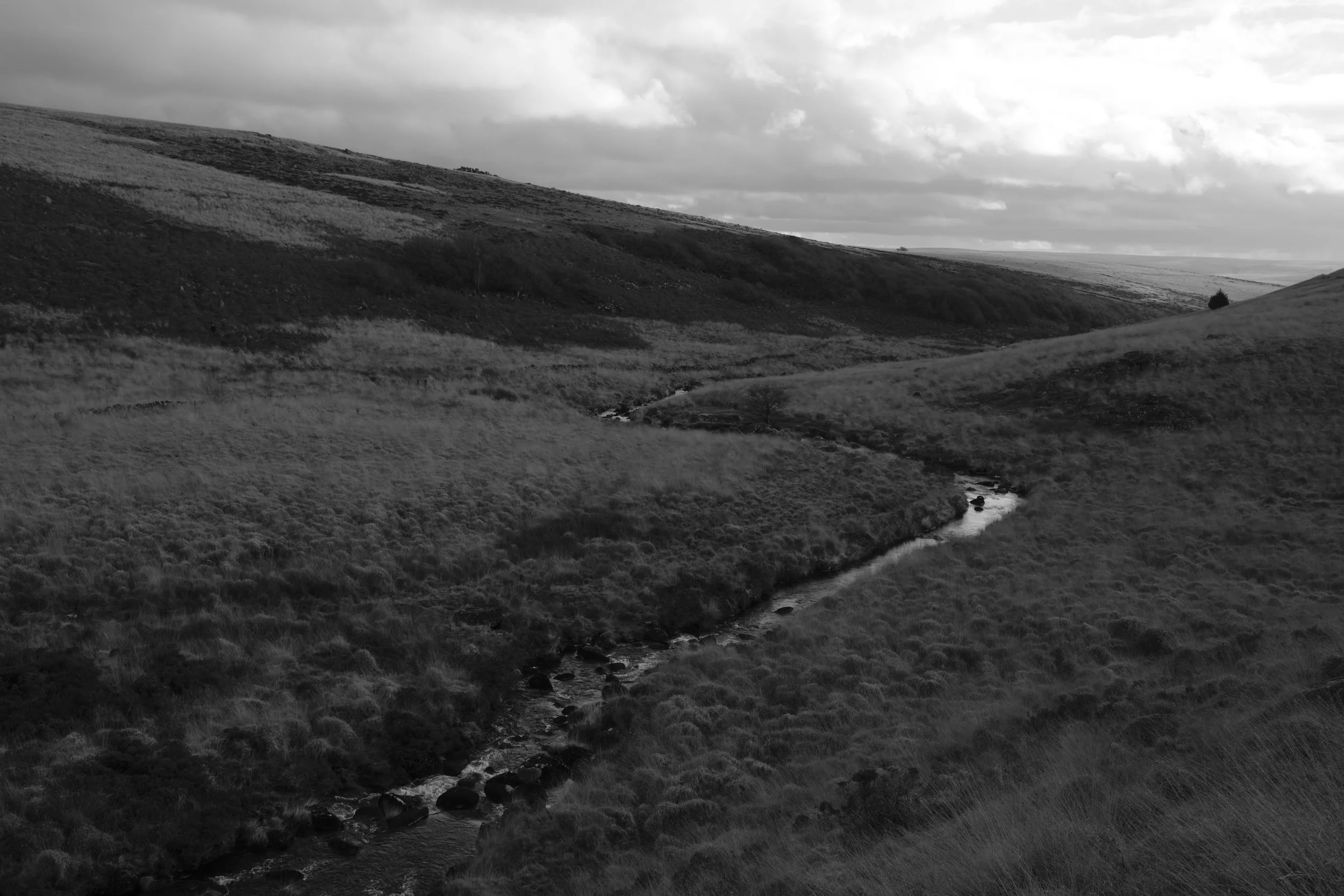Black and white landscape of a serene moor, with a winding stream cutting through grassy hills under a cloudy sky, evoking a tranquil, timeless mood.