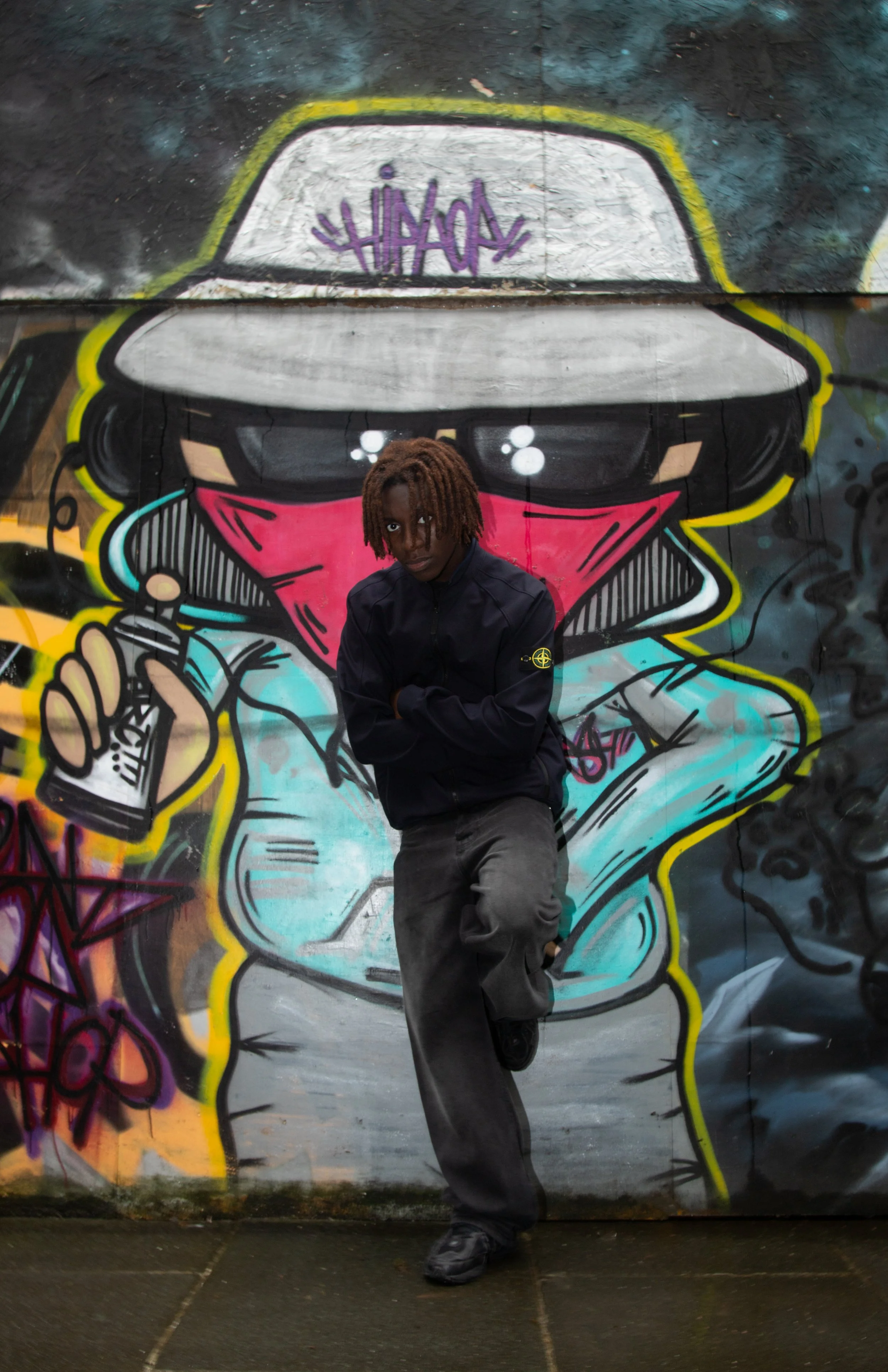 A person in black clothing poses in front of colorful graffiti featuring a masked character with a spray can. The scene conveys urban and artistic energy.