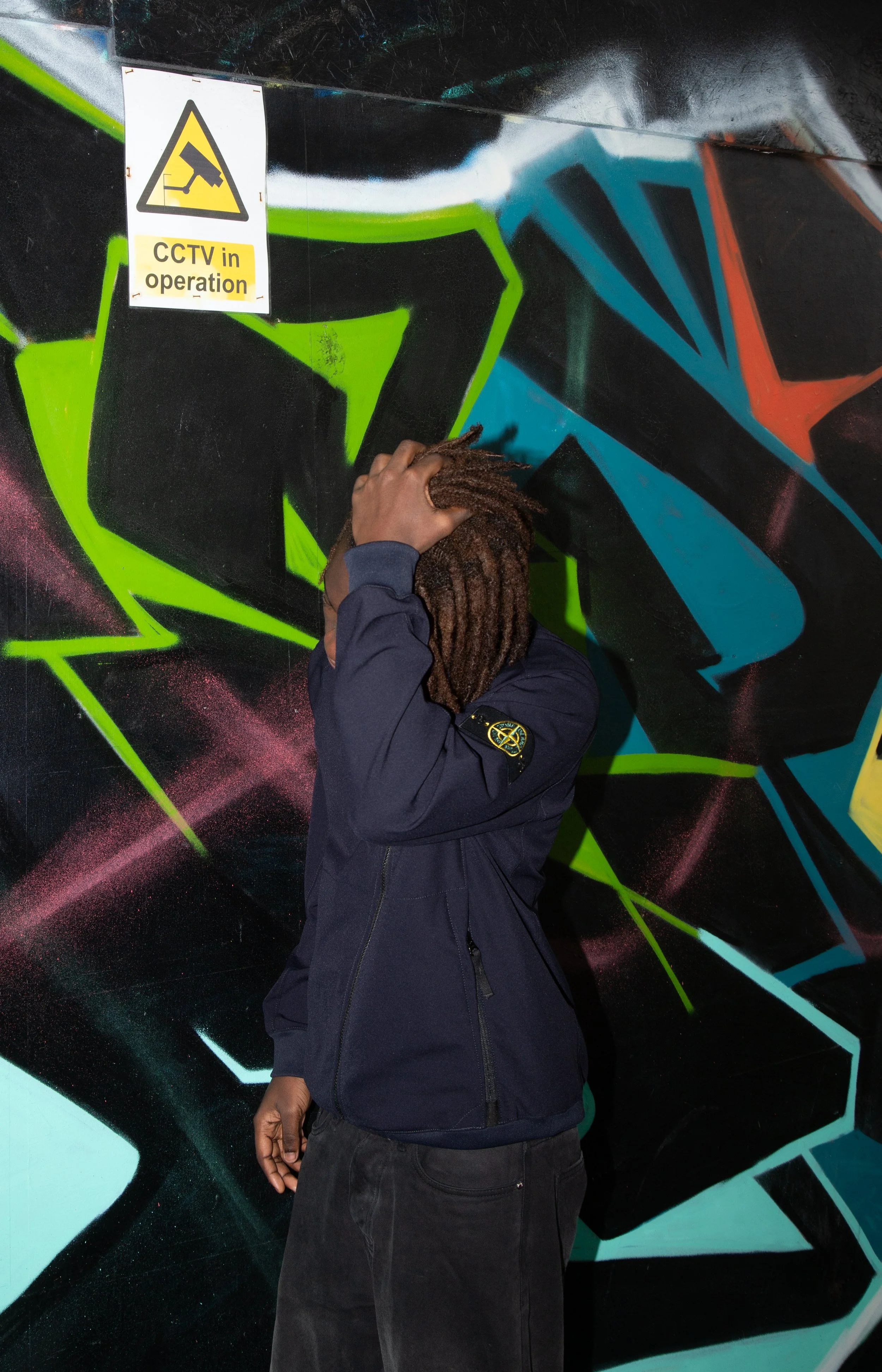A person with dreadlocks stands against vibrant graffiti, holding their head in a navy jacket. Above, a "CCTV in operation" sign is visible.