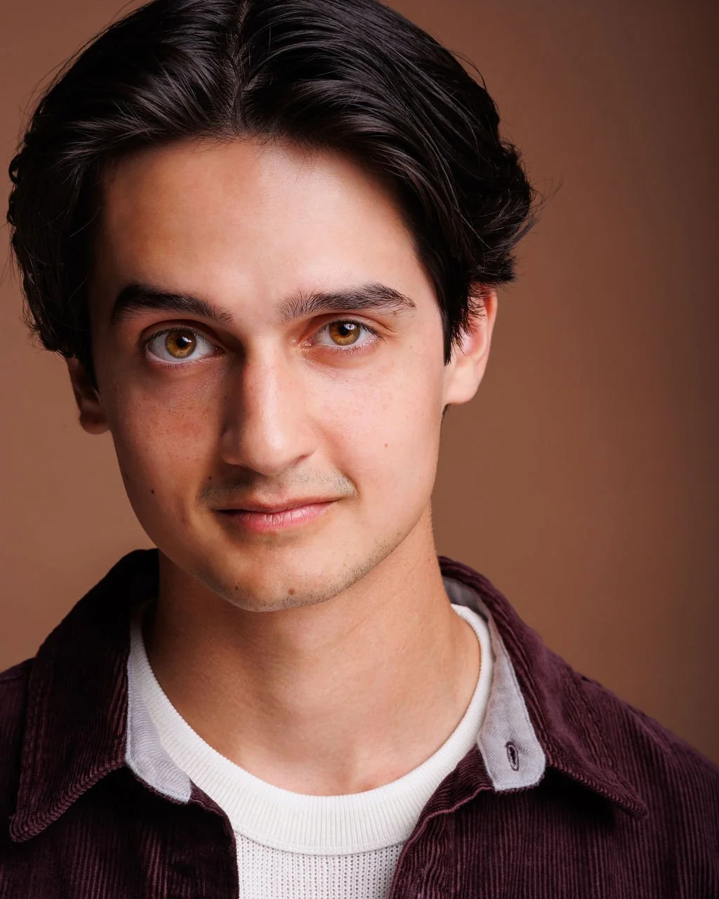 A young man with dark hair, hazel eyes, and light skin, smiling softly, wearing a white t-shirt under a burgundy jacket.