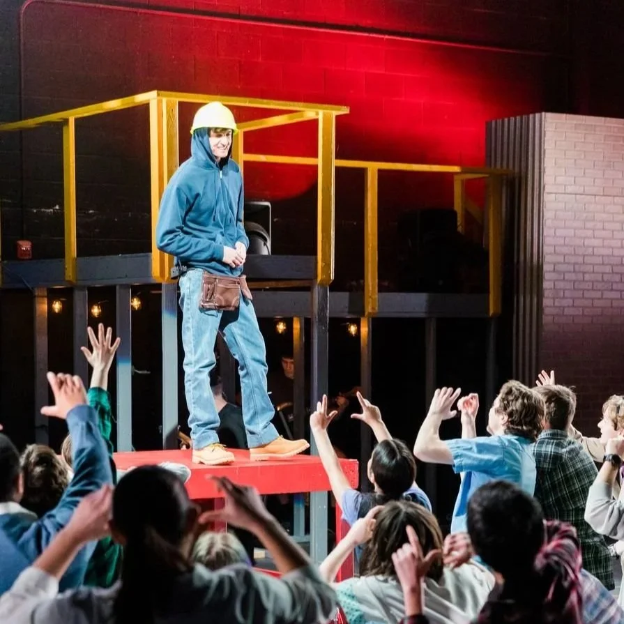 A man wearing a yellow hard hat, blue hoodie, jeans, and work boots stands on a stage in front of a group of children. The children are raising their hands and appear to be engaging with the man. The background is industrial style with dark walls and a red lighting highlight.