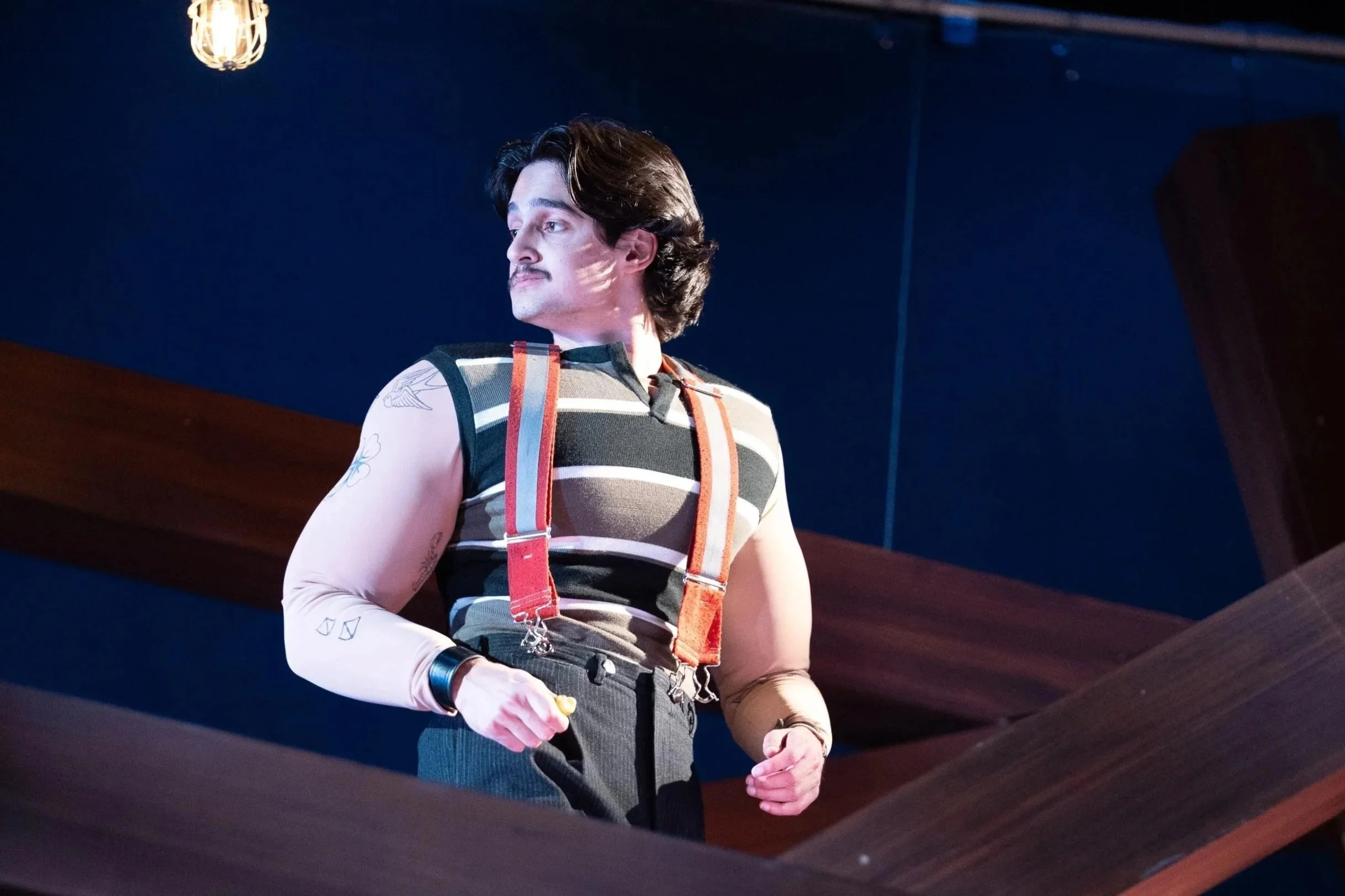 A man standing indoors, wearing a sleeveless striped shirt, suspenders, and pink long-sleeve shirt with tattoos on his arms, looking to the left.