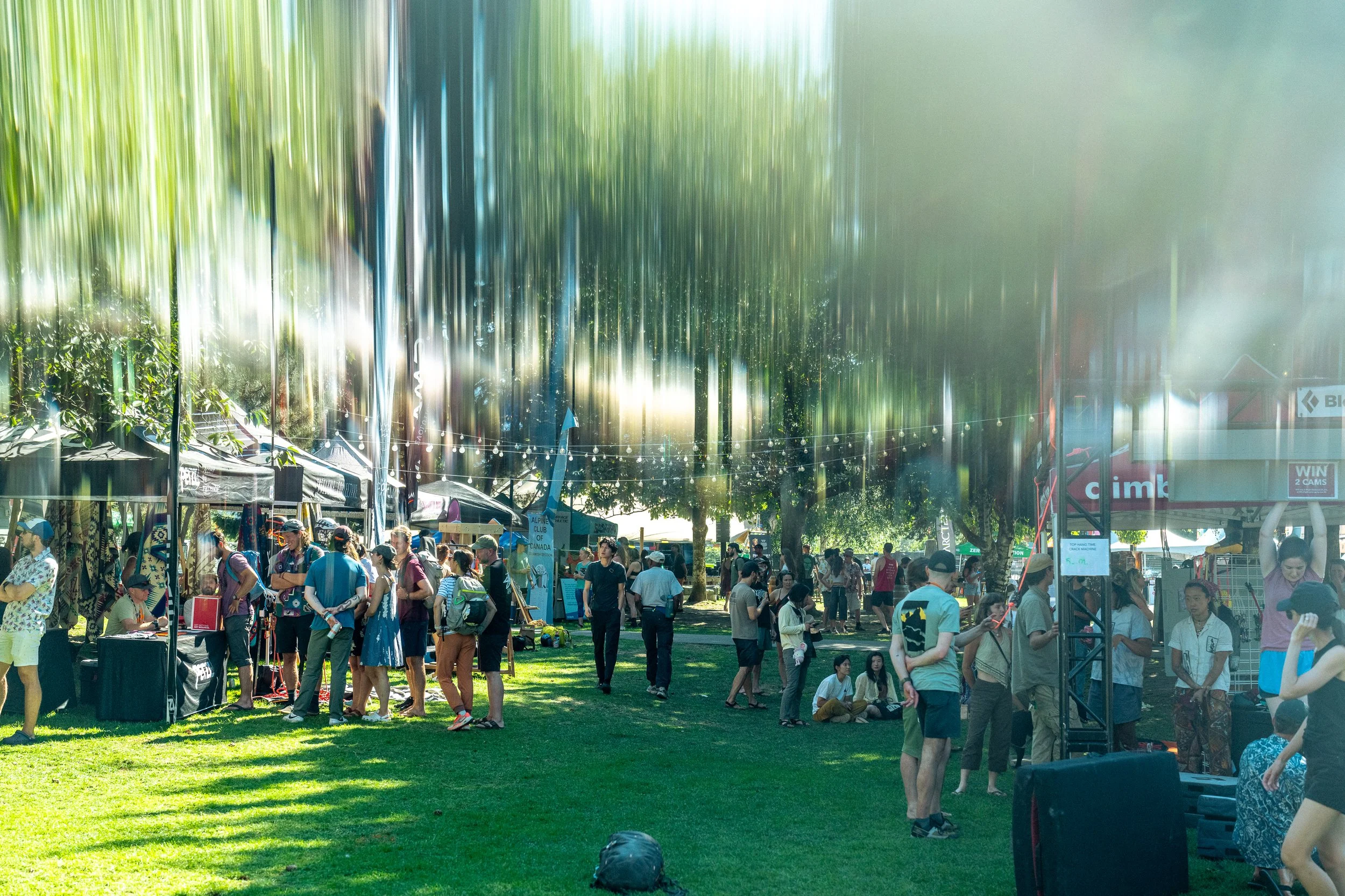 At an outdoor fair or market, people walk and browse vendor booths among trees with string lights.