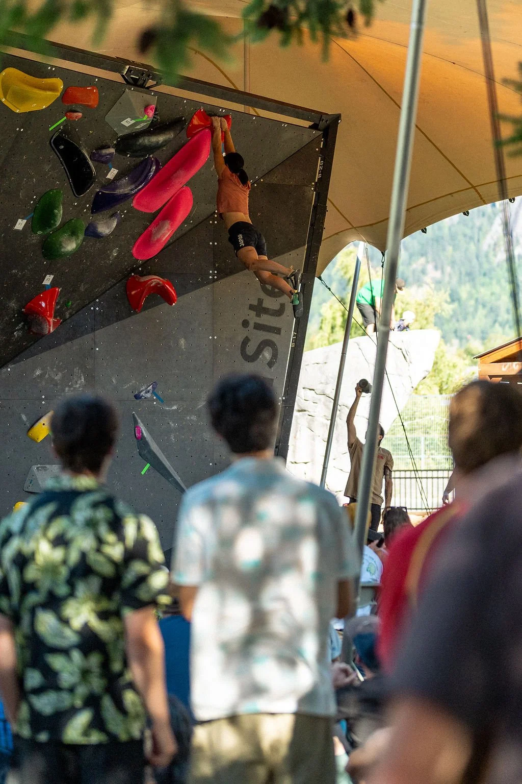 A person is climbing an indoor bouldering wall under a large yellow canopy with spectators watching.