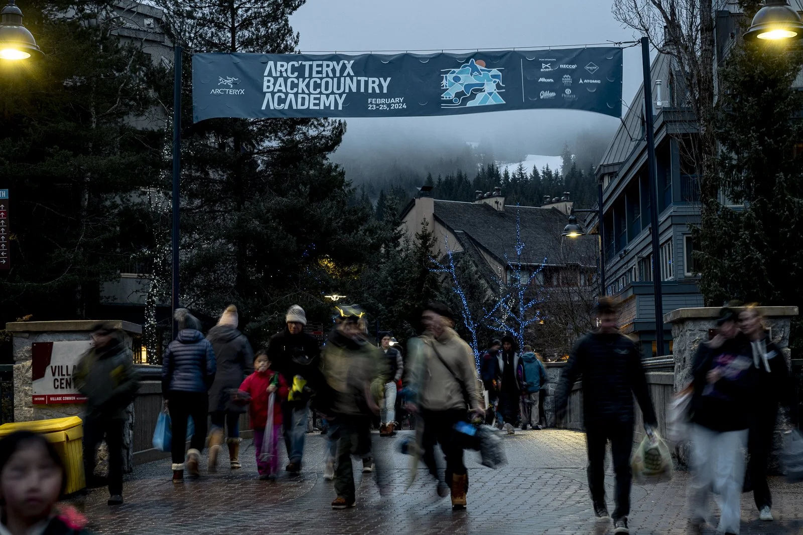 People walking through a village with a banner overhead promoting the Arc'teryx Backcountry Academy event from February 23-25, 2024, in a town surrounded by trees and buildings, with some blue and yellow lights and snow on the mountain in the backgro
