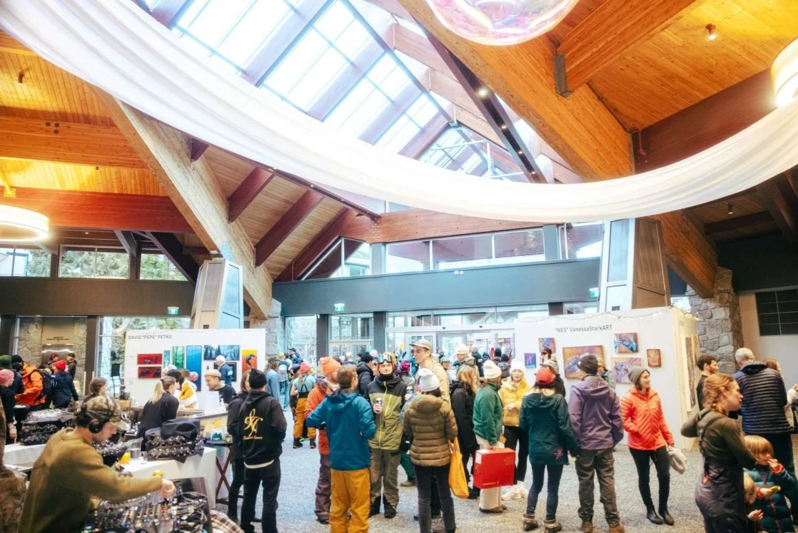 Indoor art exhibition filled with people viewing artwork and browsing booths in a spacious, modern venue with high wooden ceilings and large skylights.