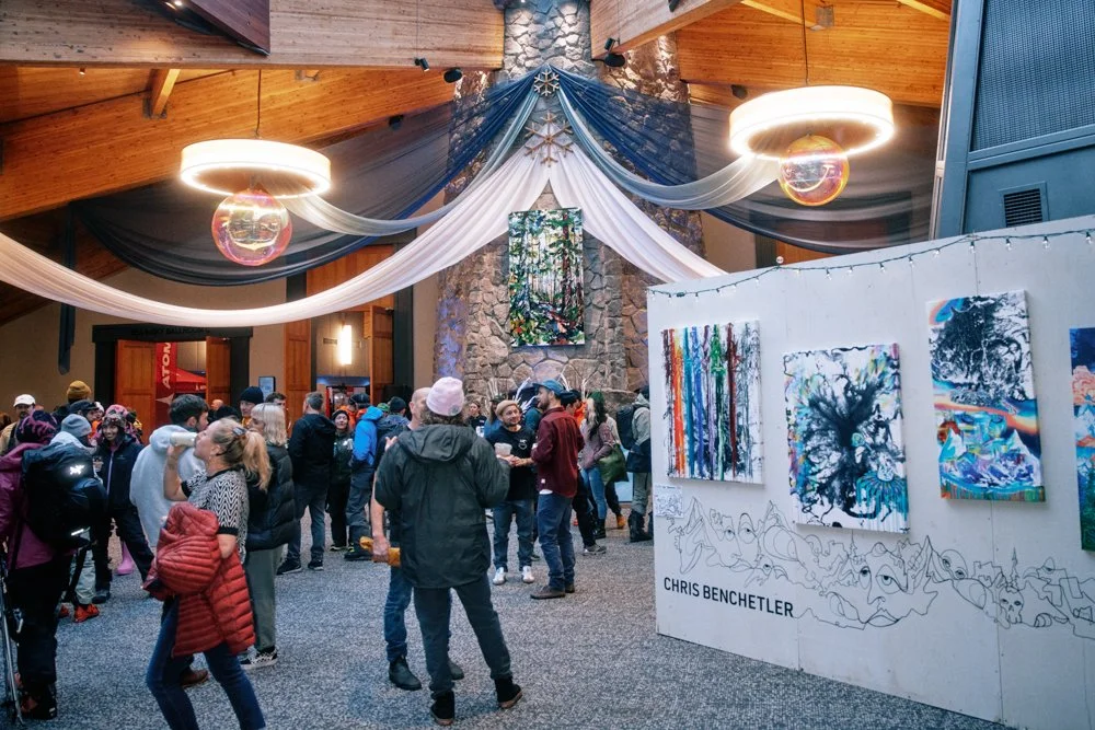 People attending an art exhibition in a large, wood-ceiling building with hanging light fixtures and a stone fireplace. Artworks by Chris Benchtler are displayed on a white wall in the foreground.