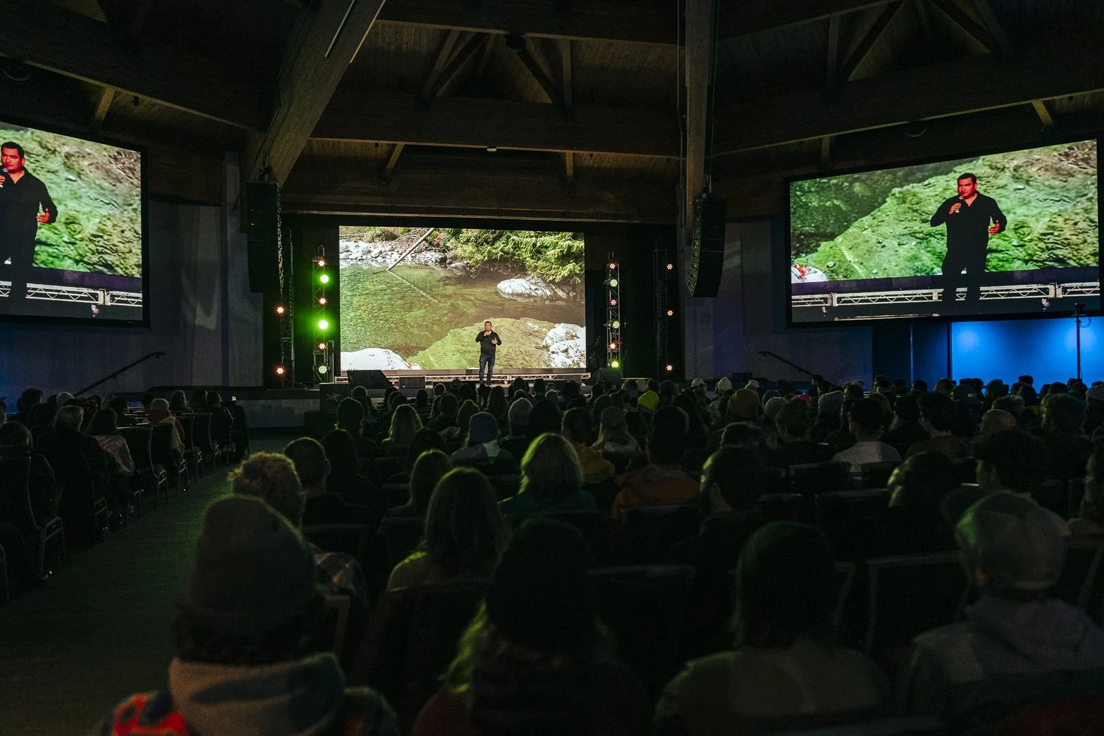 A black speaker on a stage with a presentation screen showing an outdoor scene of a river and rocks. A man in dark clothing is standing on the stage speaking to an audience in a dark room.