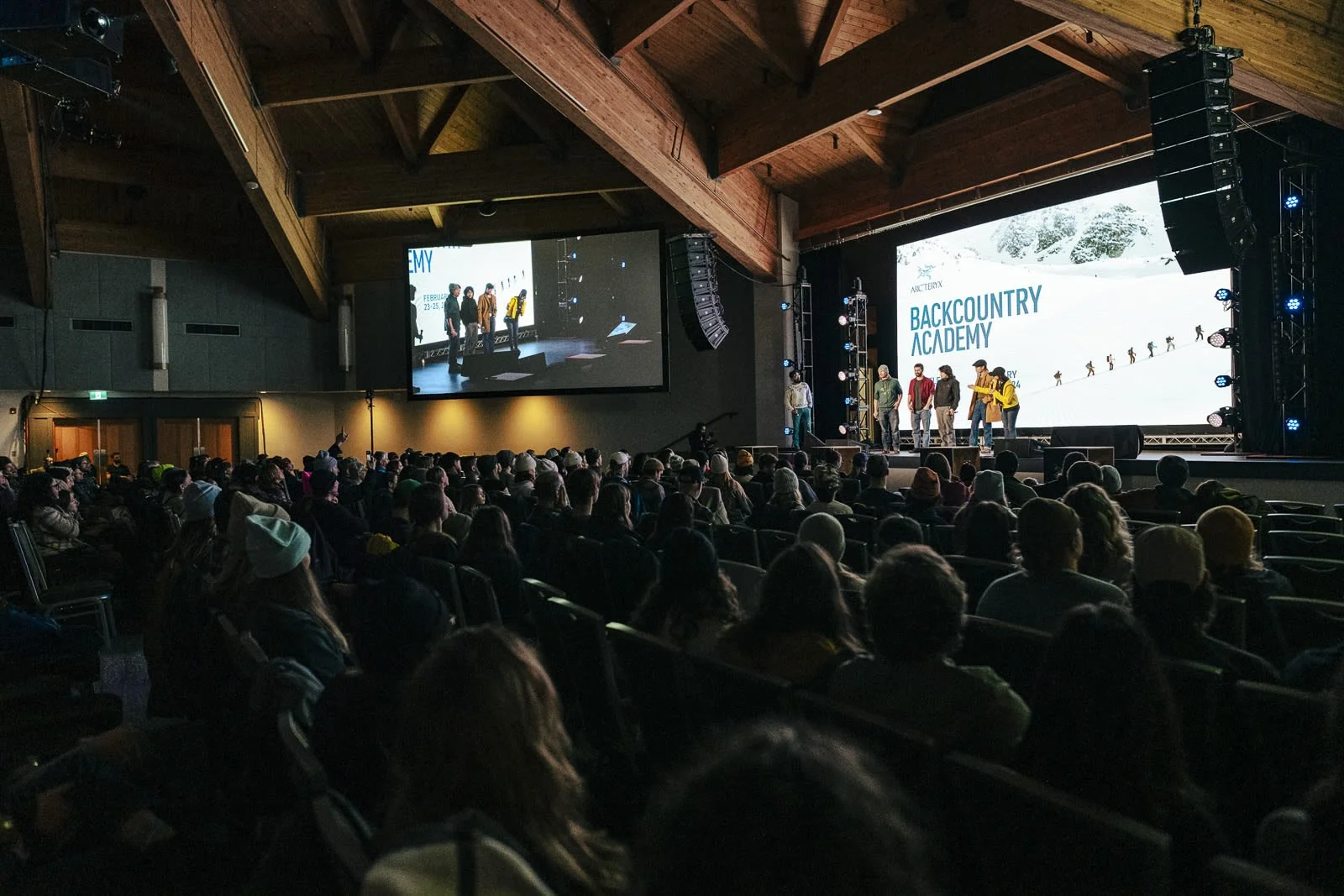 Audience watching a presentation on stage about backcountry skiing at the Backcountry Academy event with a large screen displaying the event name and visuals of skiers.