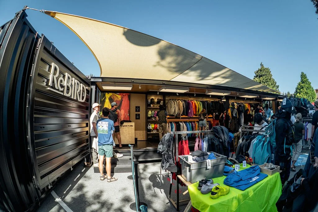 Outdoor clothing and gear shop at a market, with racks of jackets and hats, and customers browsing and shopping.