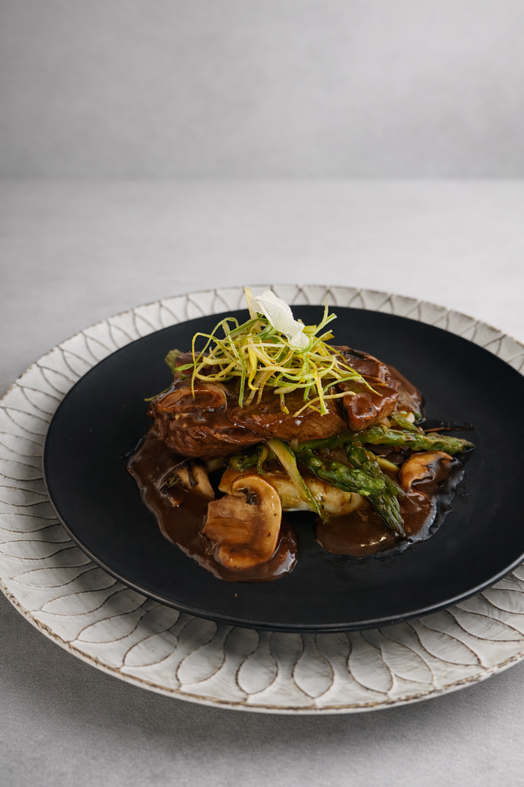 Plato de carne con salsa oscura, servido con champiñones, espárragos y guarnición de vegetales, decorado con cebollín o cebolleta.