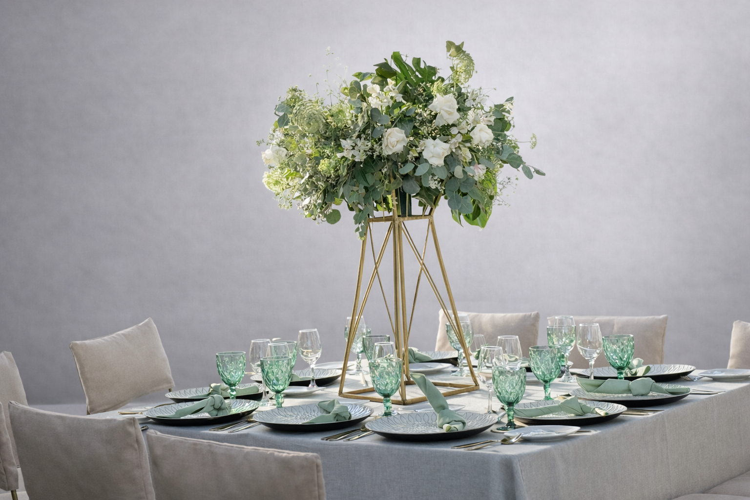 Mesa decorada con vajilla, copas de vino y agua, cubiertos y servilletas, con un centro de flores blancas y verdes en un soporte dorado.