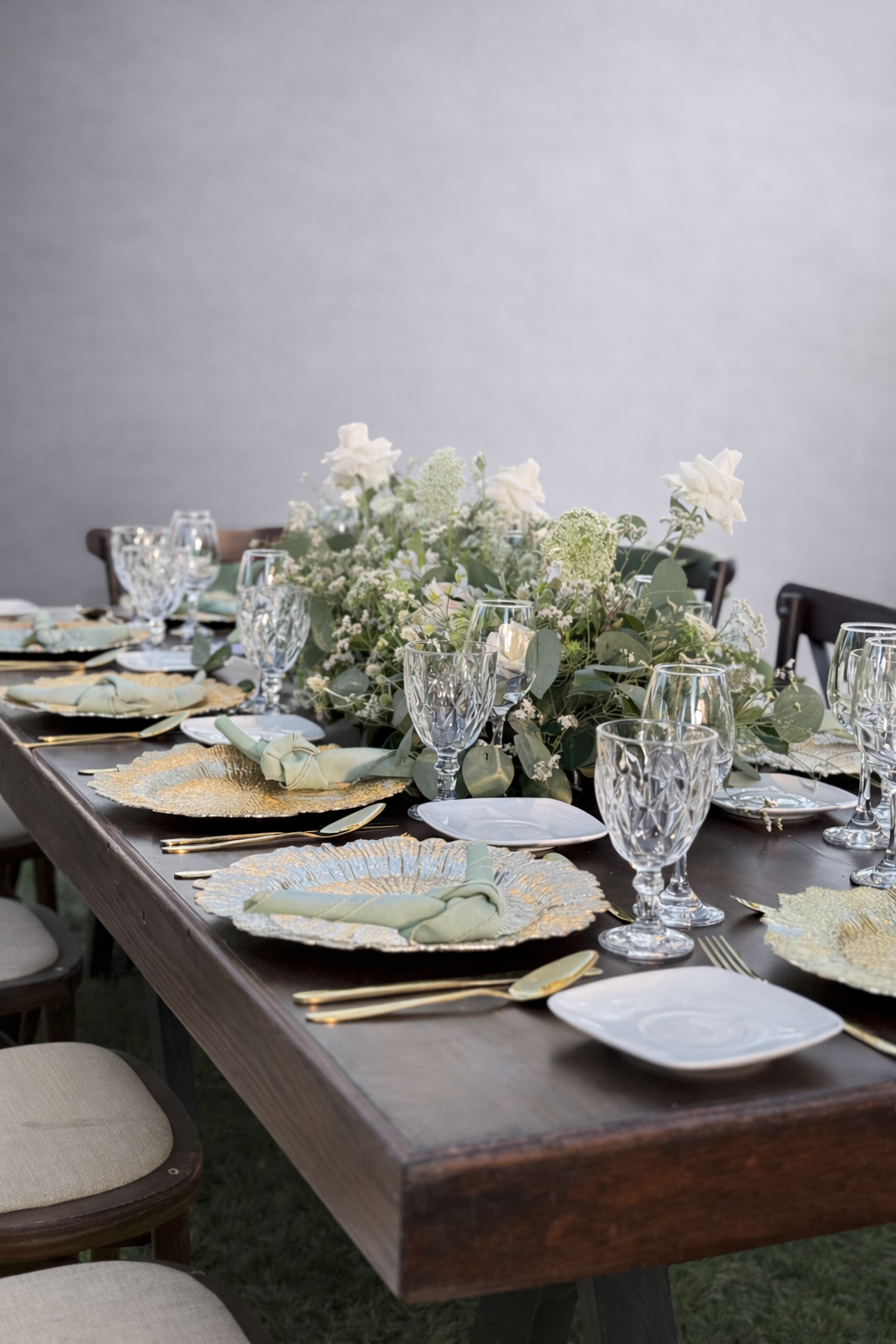 Mesa de comedor decorada para un evento con un centro de flores blancas y verde, platos dorados, copas, cubiertos y servilletas en colores pastel.