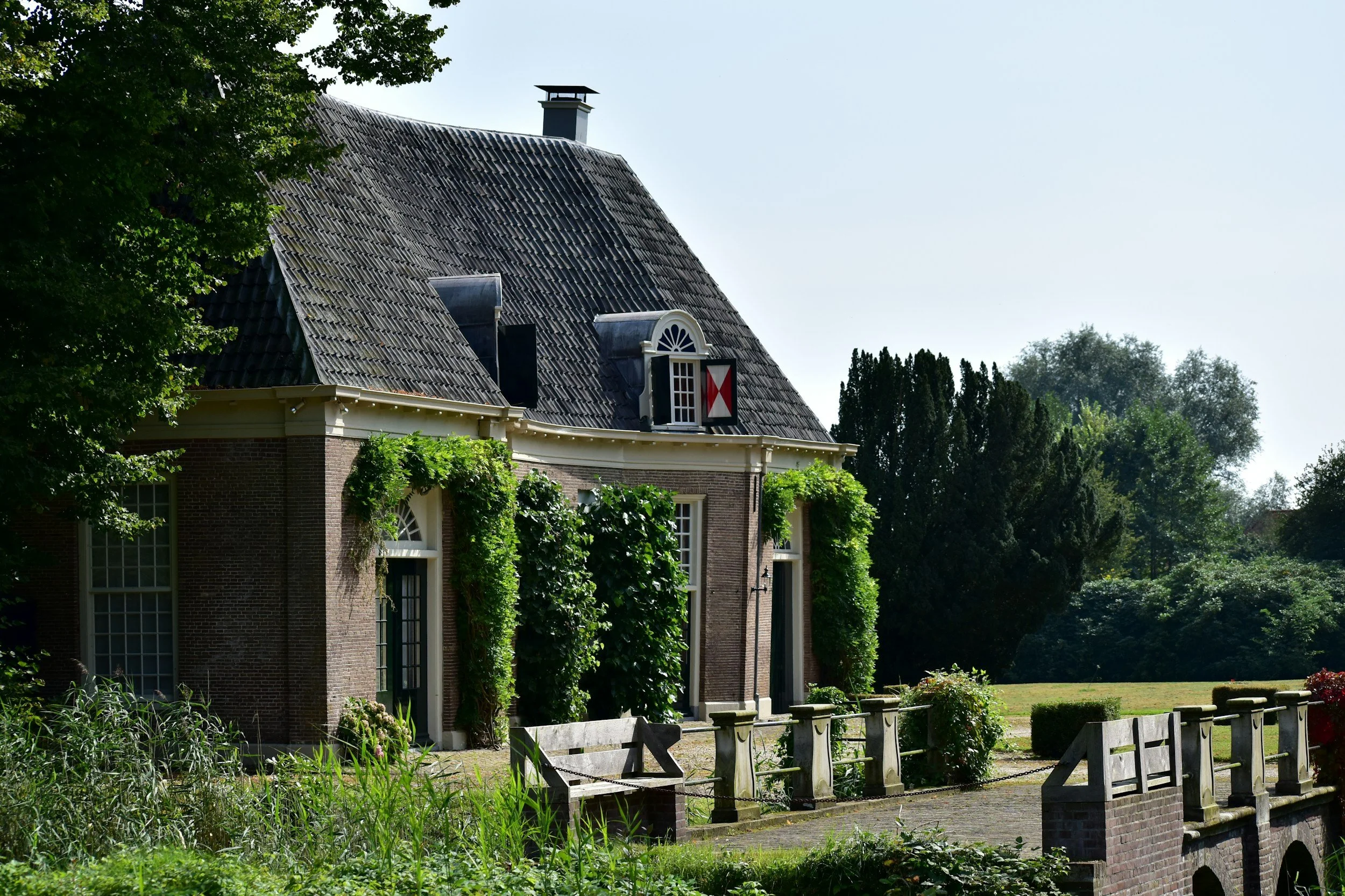 A brick house with a steep, tiled roof, surrounded by green vegetation and trees, with a small stone bridge in the foreground.