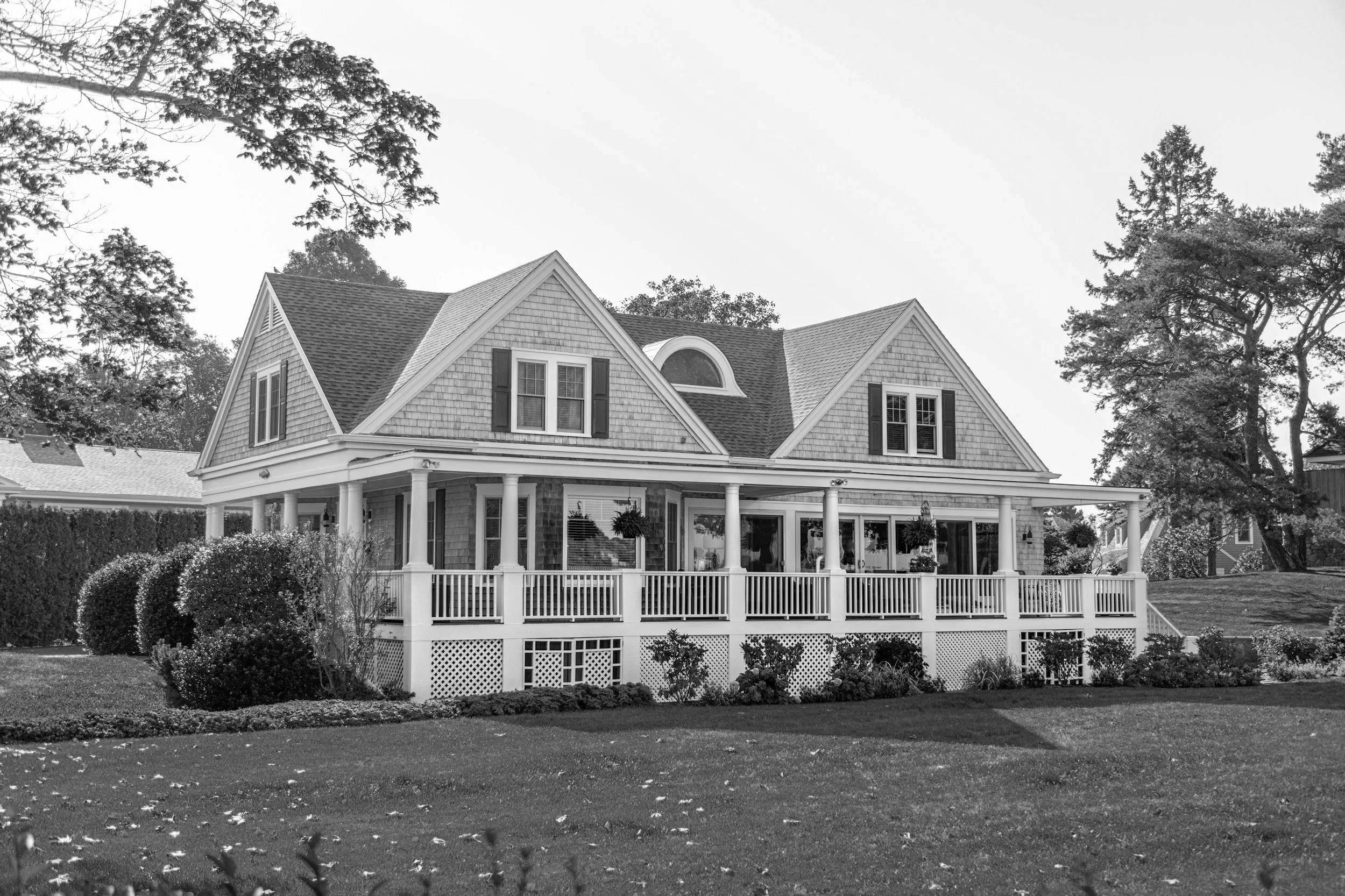 A large house with a wrap-around porch, multiple gabled roofs, and a dormer window, surrounded by a well-maintained lawn and trees, in black and white.