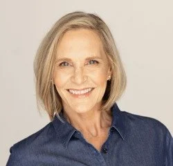 A woman with shoulder-length gray hair smiling, wearing a denim shirt, against a neutral background.