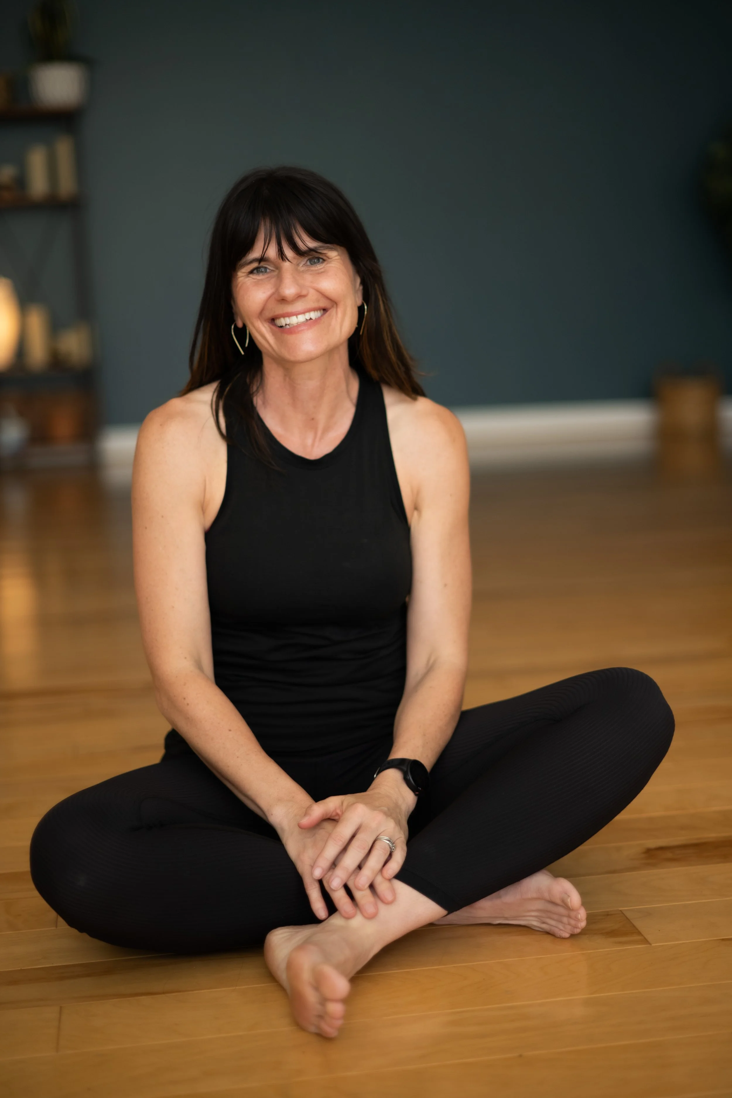 Woman sitting cross-legged on a wooden floor, smiling, wearing a black sleeveless top, black leggings, and a watch, in a room with a dark blue wall and a shelf in the background.