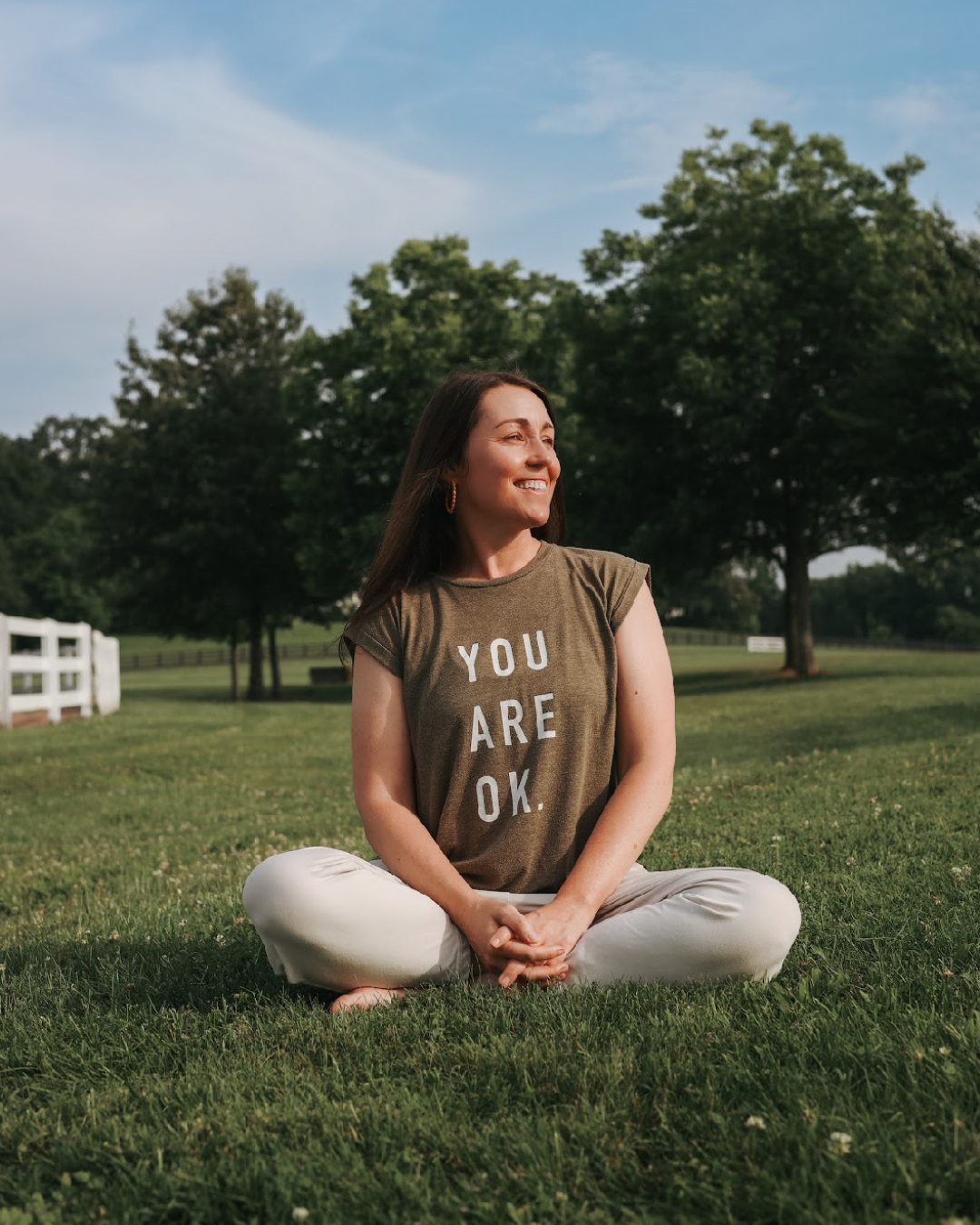A woman sitting cross-legged on grass in a park, smiling, wearing a t-shirt that says "You Are OK", with trees and a blue sky in the background.