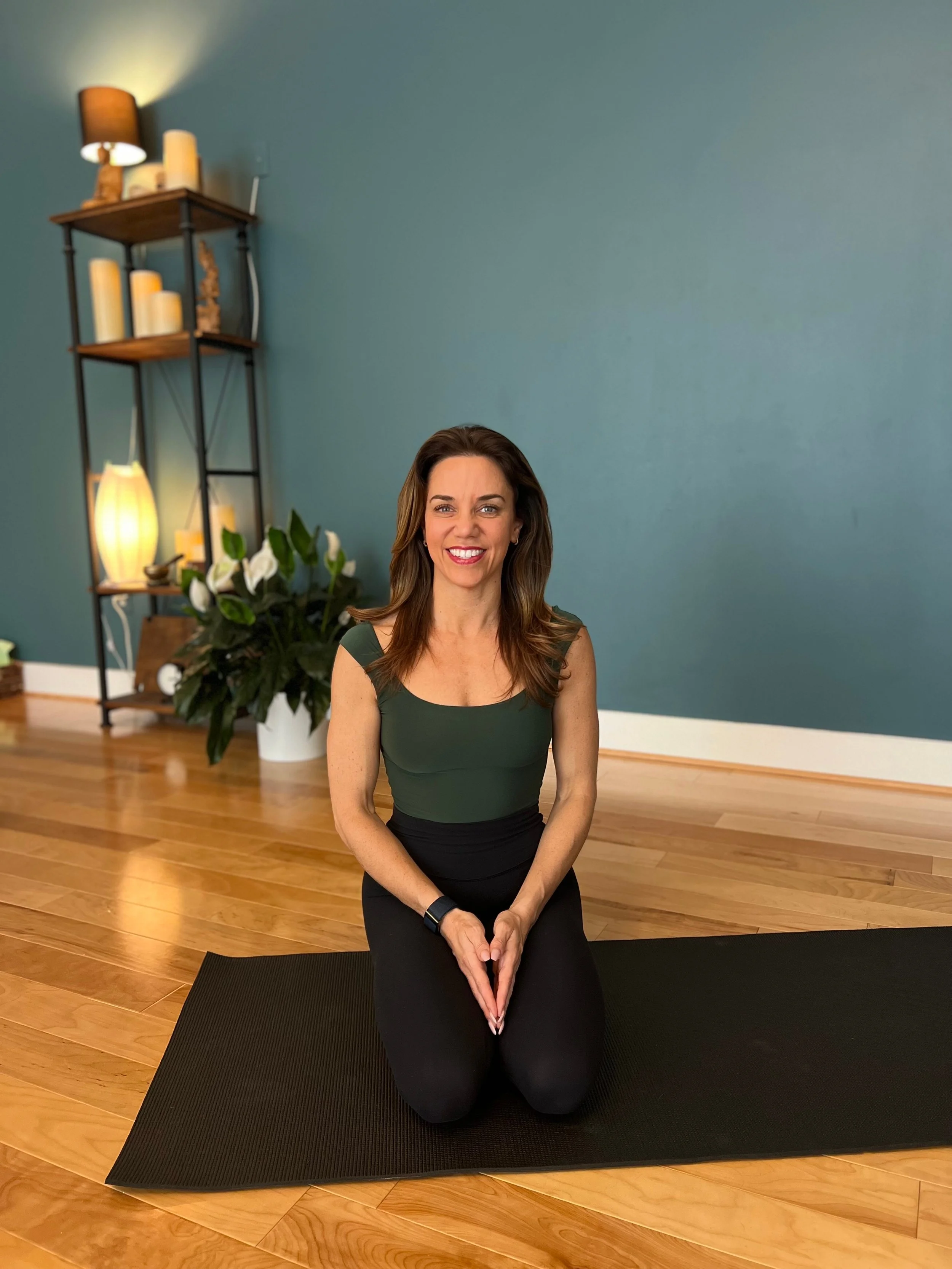 A woman with long brown hair, wearing a dark green sleeveless top and black pants, is kneeling on a black yoga mat in a room with wooden floors and teal walls. She is smiling and holding her hands together in front of her. In the background, there is a shelf with candles and a lamp, and a large potted plant.