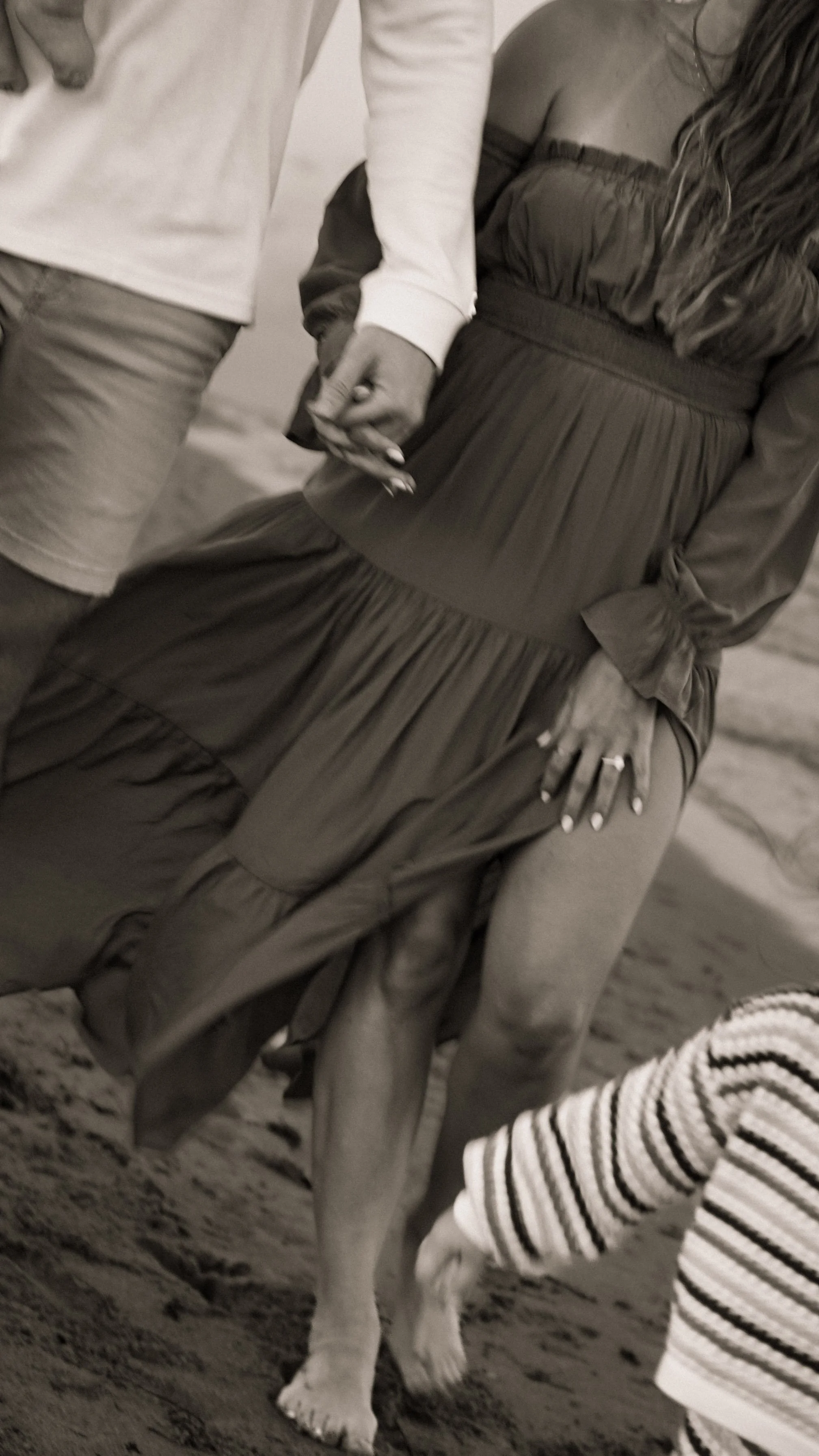 A woman with dark hair is hugging a man with a shaved head at the beach, smiling happily.
