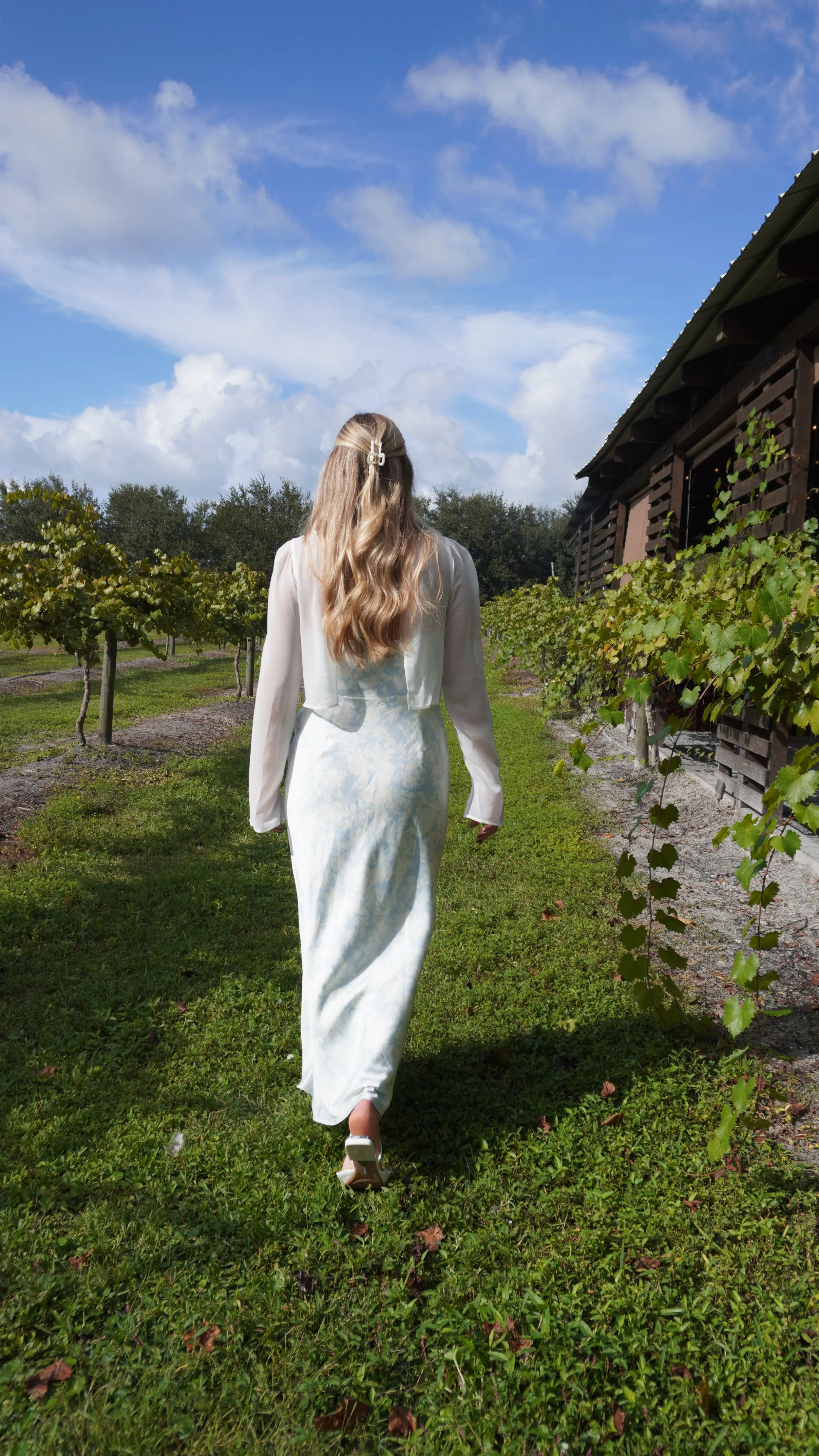 A bride and groom are standing outdoors near a white building, with lush green trees in the background. The bride is holding a bouquet of white calla lilies and is wearing a white sleeveless wedding gown. The groom is dressed in a dark suit with a white shirt. They are looking at each other, smiling.