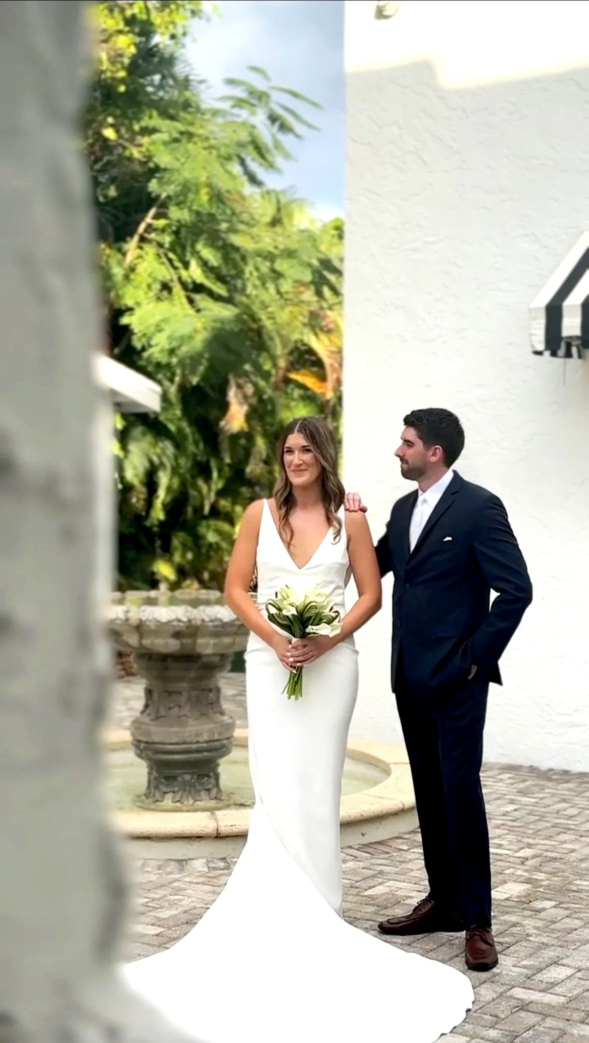 A bride and groom are standing outdoors near a white building, with lush green trees in the background. The bride is holding a bouquet of white calla lilies and is wearing a white sleeveless wedding gown. The groom is dressed in a dark suit with a white shirt. They are looking at each other, smiling.