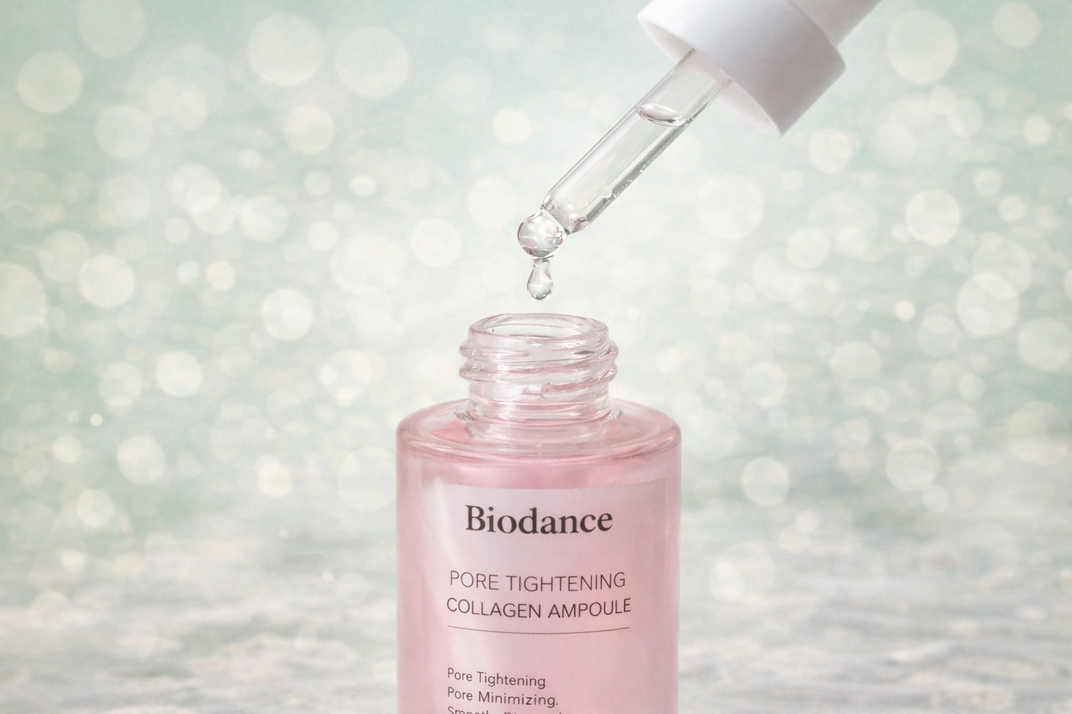 A pink bottle of Biodance pore tightening collagen ampoule with a glass dropper above the open bottle, with a blurred light blue bokeh background.