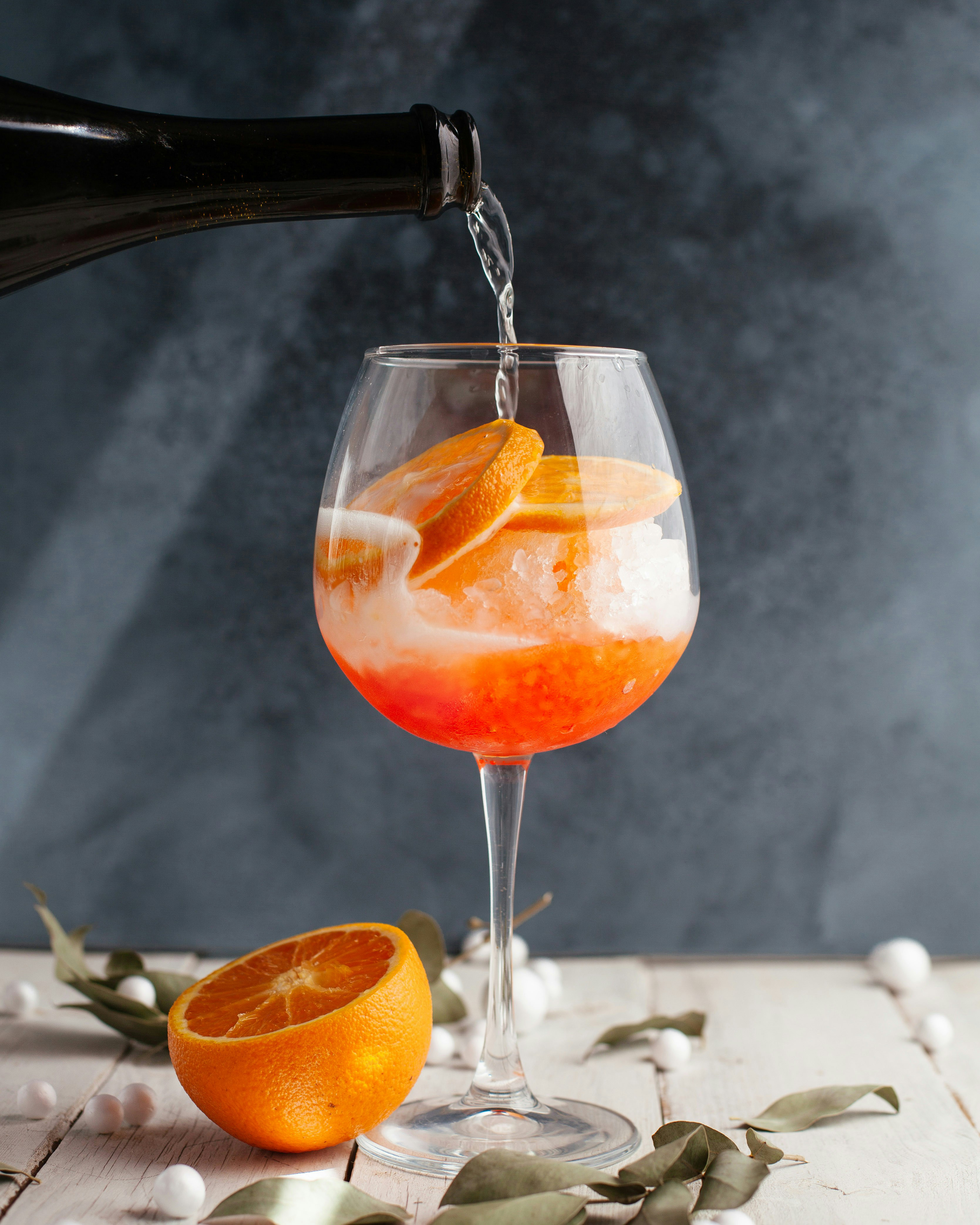 A cocktail glass with orange slices and crushed ice, being filled with sparkling water from a bottle, on a white wooden surface with an orange half and scattered leaves and small white round decorations.