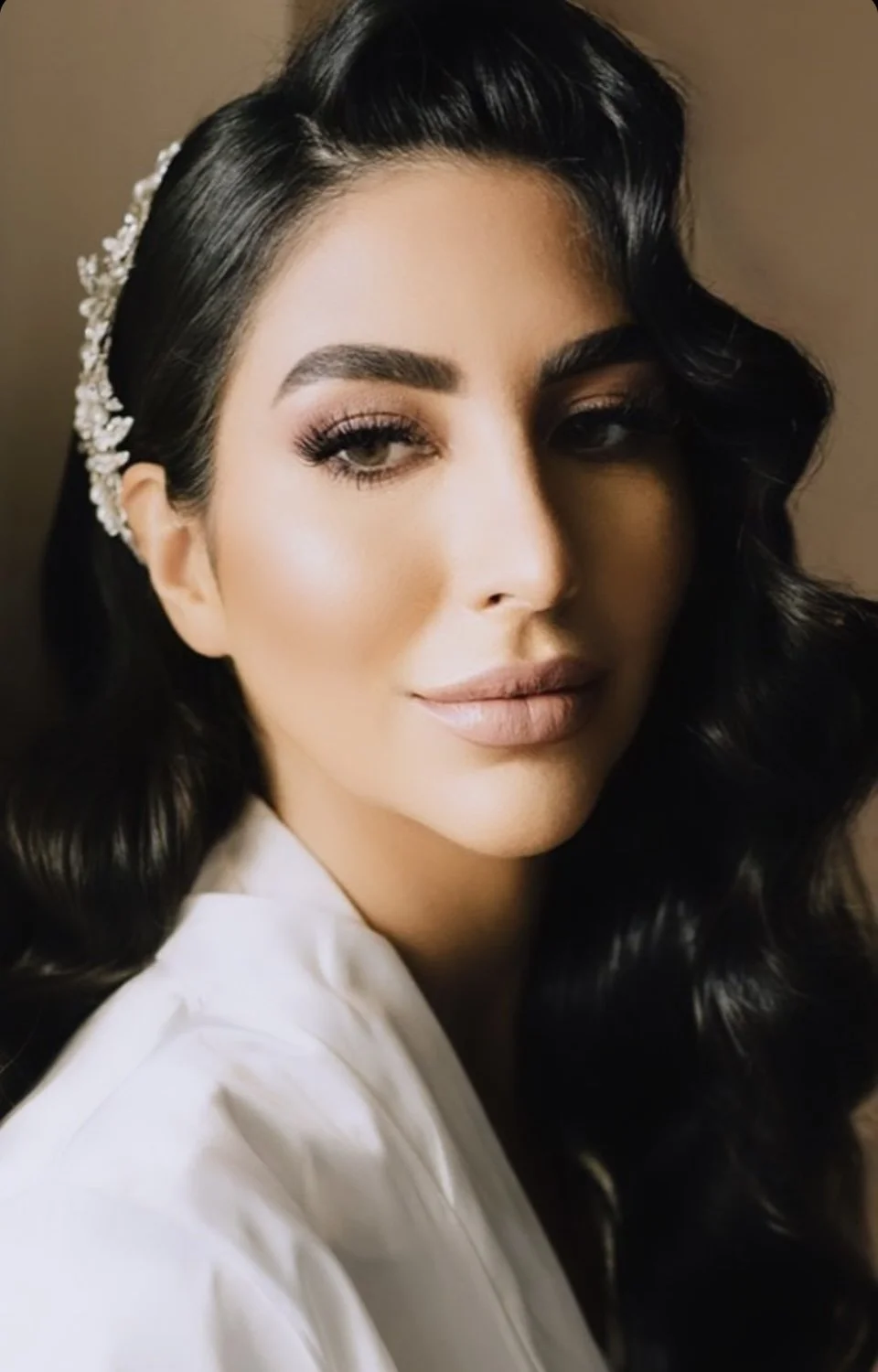 Bride with dark, Hollywood wave styled hair wearing a pearl-embellished hairpiece, makeup with defined eyebrows, long lashes, and nude lipstick, dressed in a white wedding dressing gown.