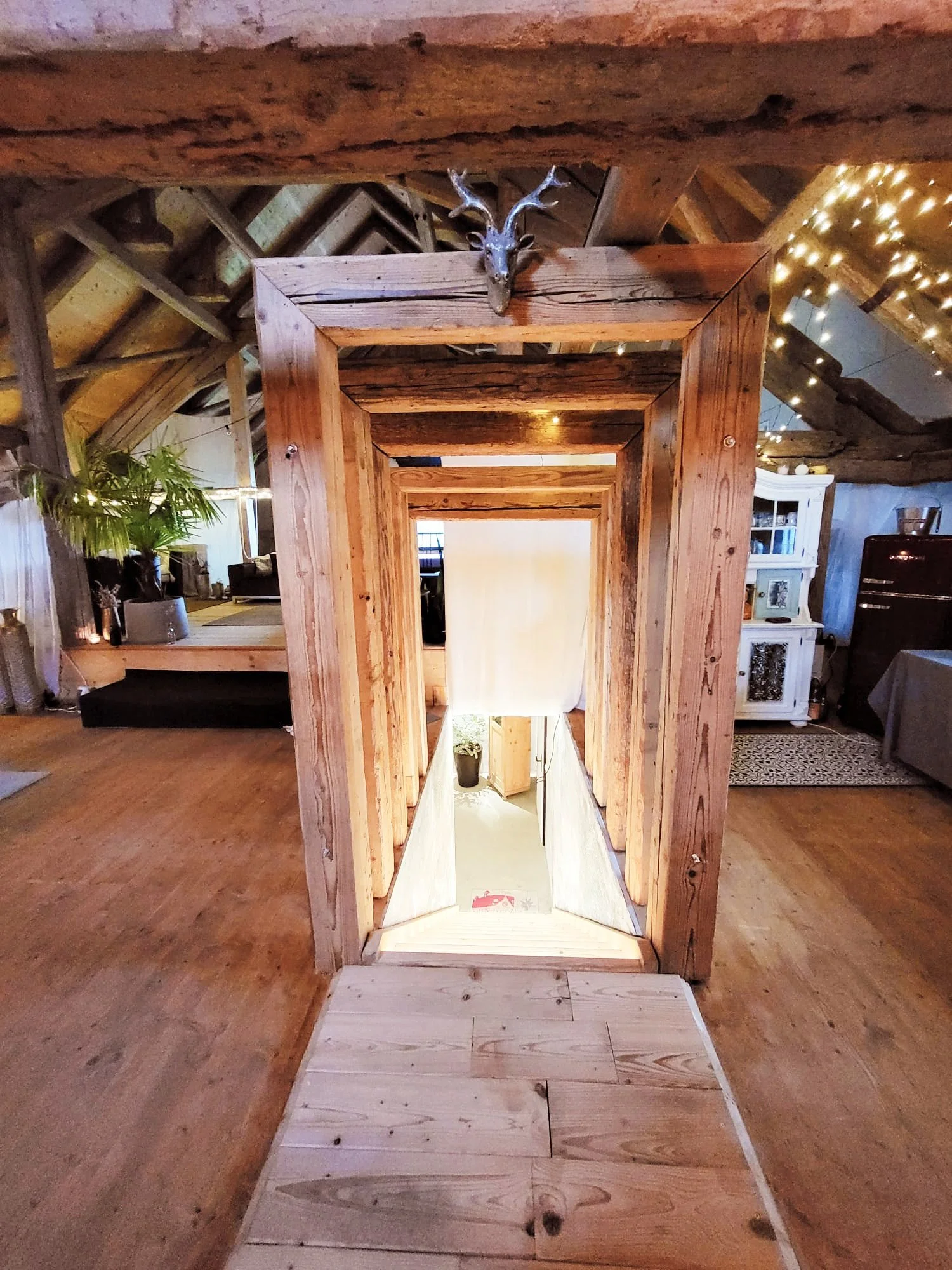 Ein Blick in einen rustikalen, offenen Dachboden mit Holzbalken. Im Zentrum führt eine kleine Holztreppe nach unten. Oben an der Wand hängt ein Hirschgeweih. Die Umgebung ist warm beleuchtet und mit Dekorationen wie Lichterketten geschmückt.
