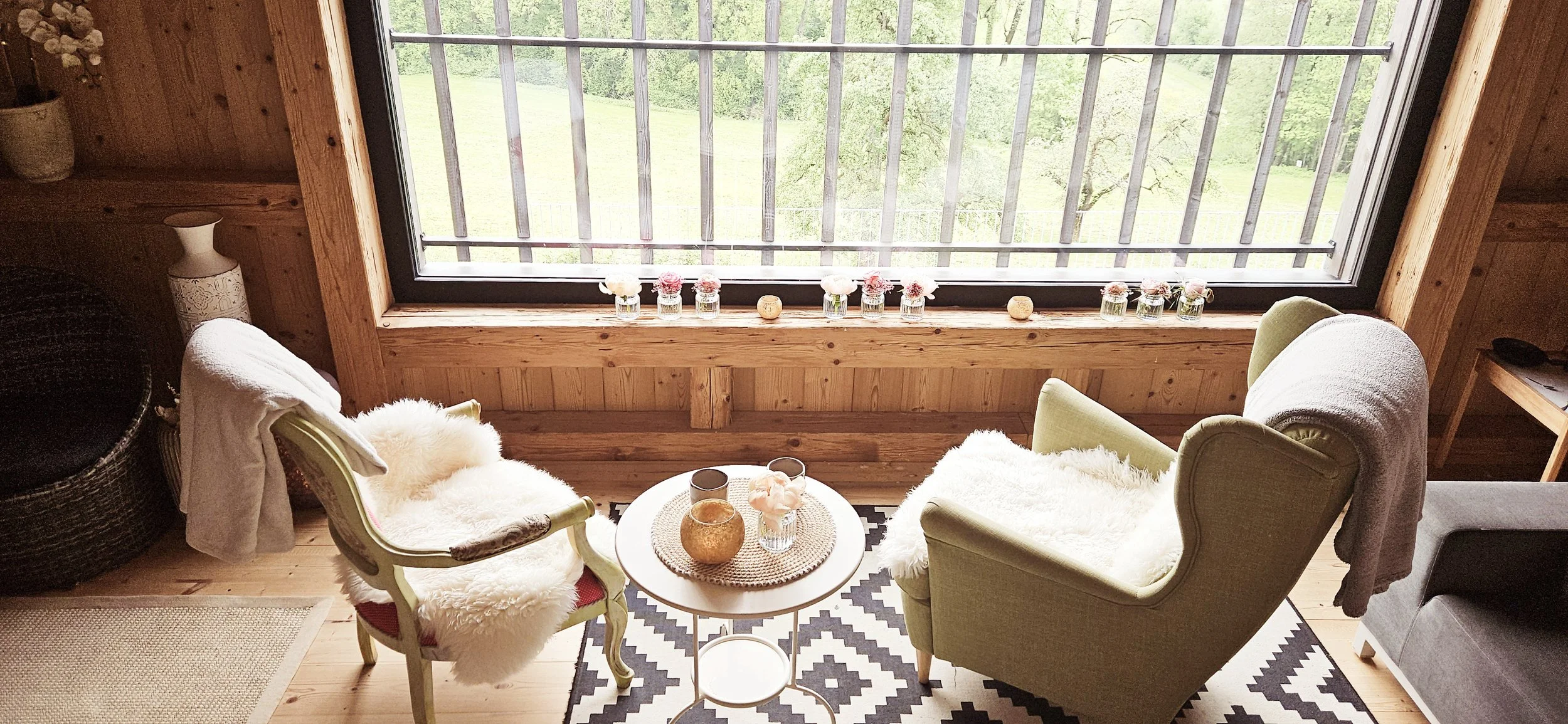Gemütliches Wohnzimmer mit zwei bequemen Sesseln, einem kleinen runden Tisch mit Dekorvasen, und einem großen Fenster mit Blick auf eine grüne Landschaft, geschmückt mit kleinen Vasen und Dekorsteinen auf der Fensterbank.