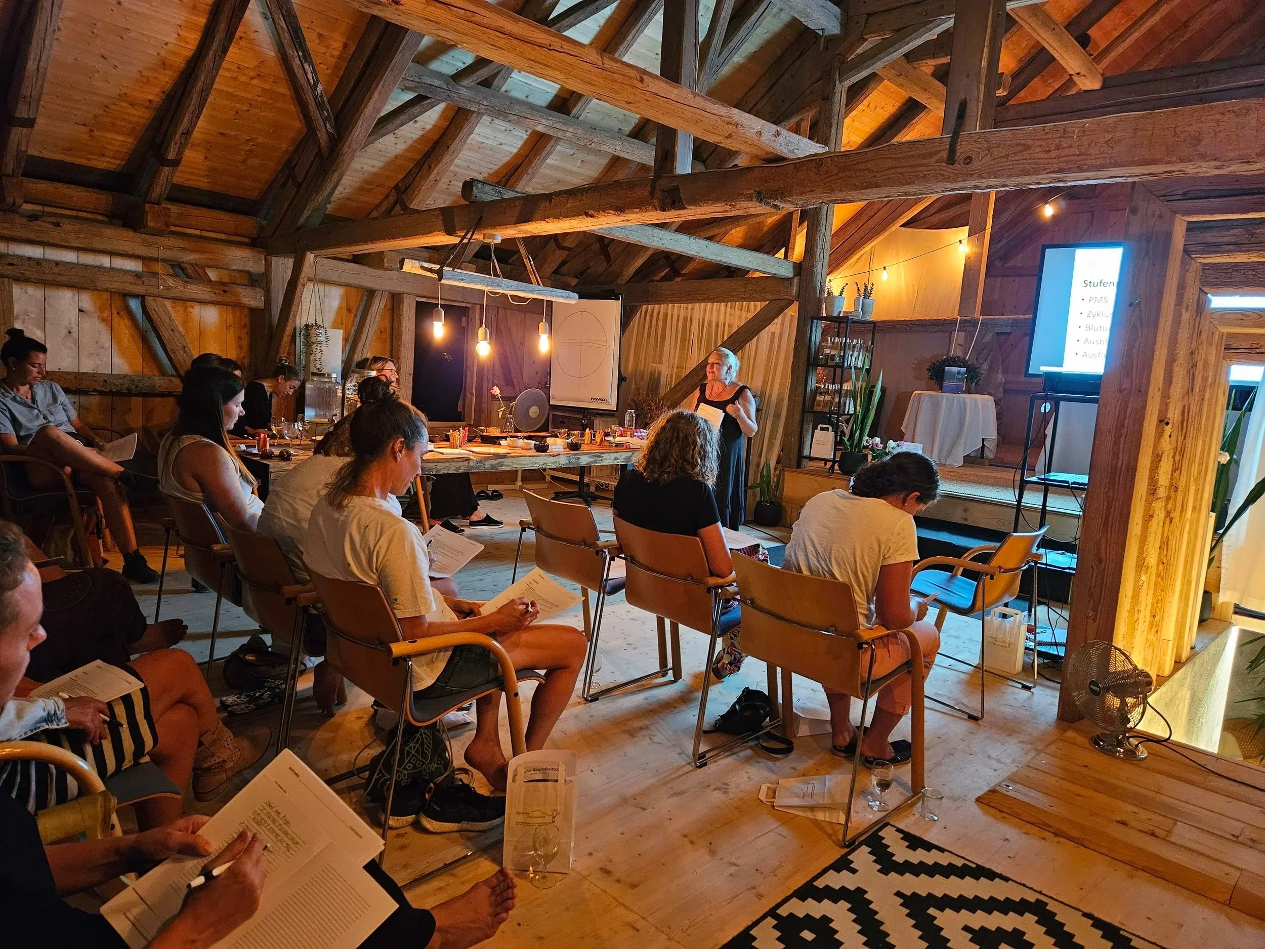 Menschen bei einem Workshop in einer wunderschönen umgebauten Scheune, die auf eine Präsentation auf der Leinwand schauen, sitzen in Stühlen, einige haben Unterlagen in der Hand.